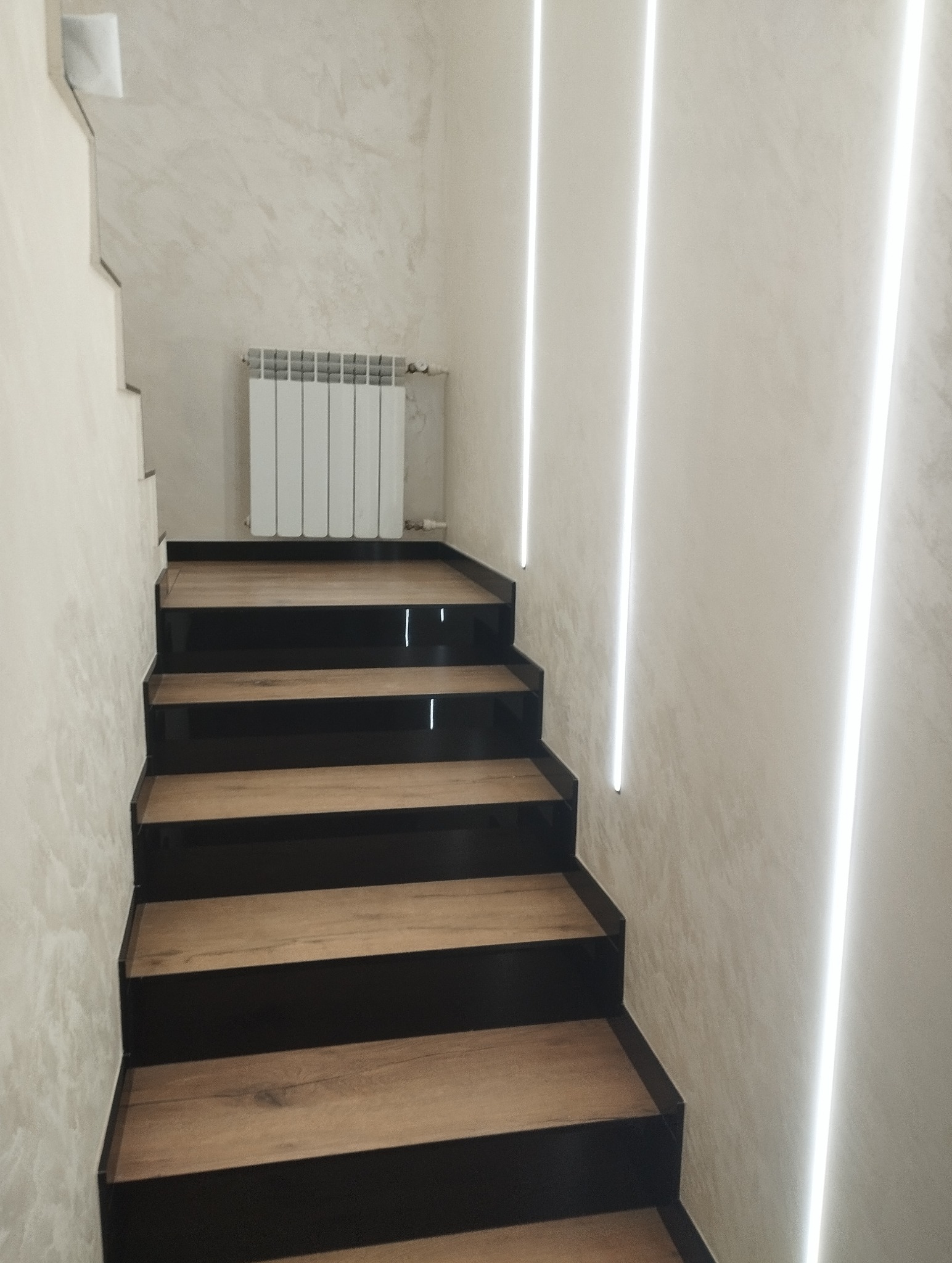 Nowoczesne schody z drewnianymi stopniami i czarnymi podstopnicami, oświetlone pionowymi pasami LED na ścianie o fakturze betonu. Grzejnik na ścianie w tle.