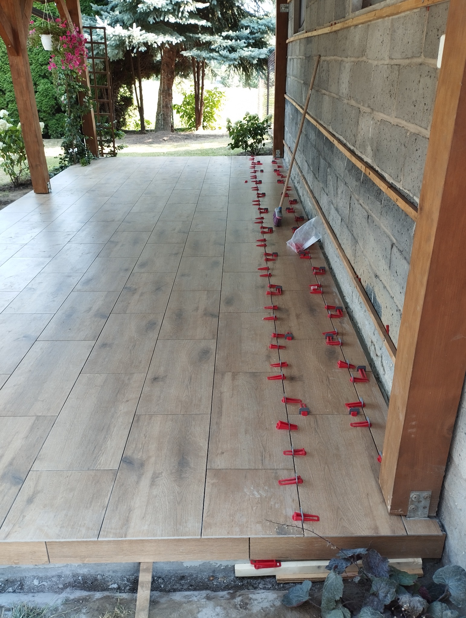 Układanie płytek imitujących drewno na zewnątrz budynku, widoczne czerwone klipsy poziomujące, tło z zielenią i elementami architektury ogrodowej.