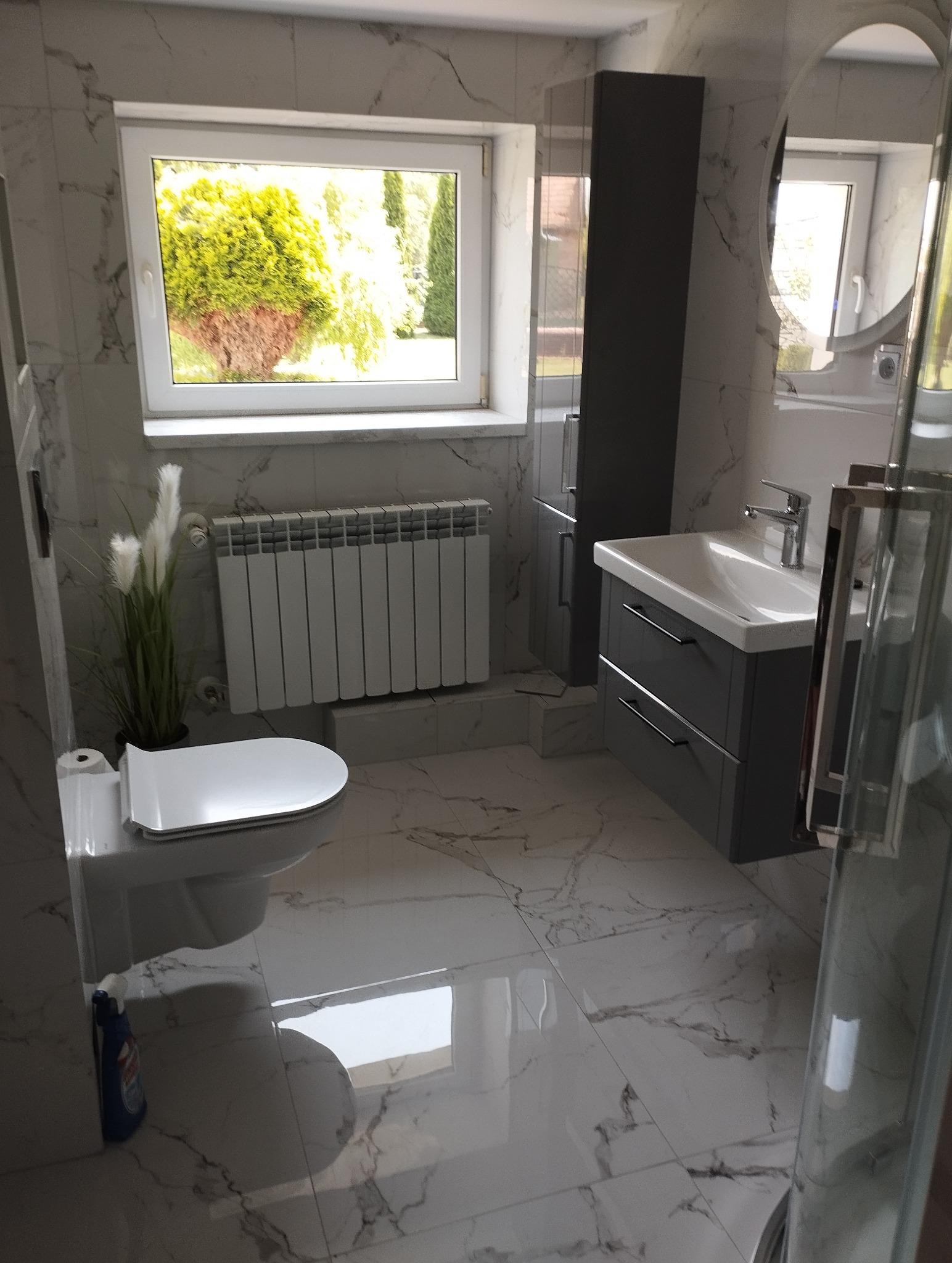 Jasna łazienka z oknem, wykończona marmurowymi płytkami. Uwagę przykuwa szara szafka pod umywalką i widok zieleni za oknem. Minimalistyczny styl.