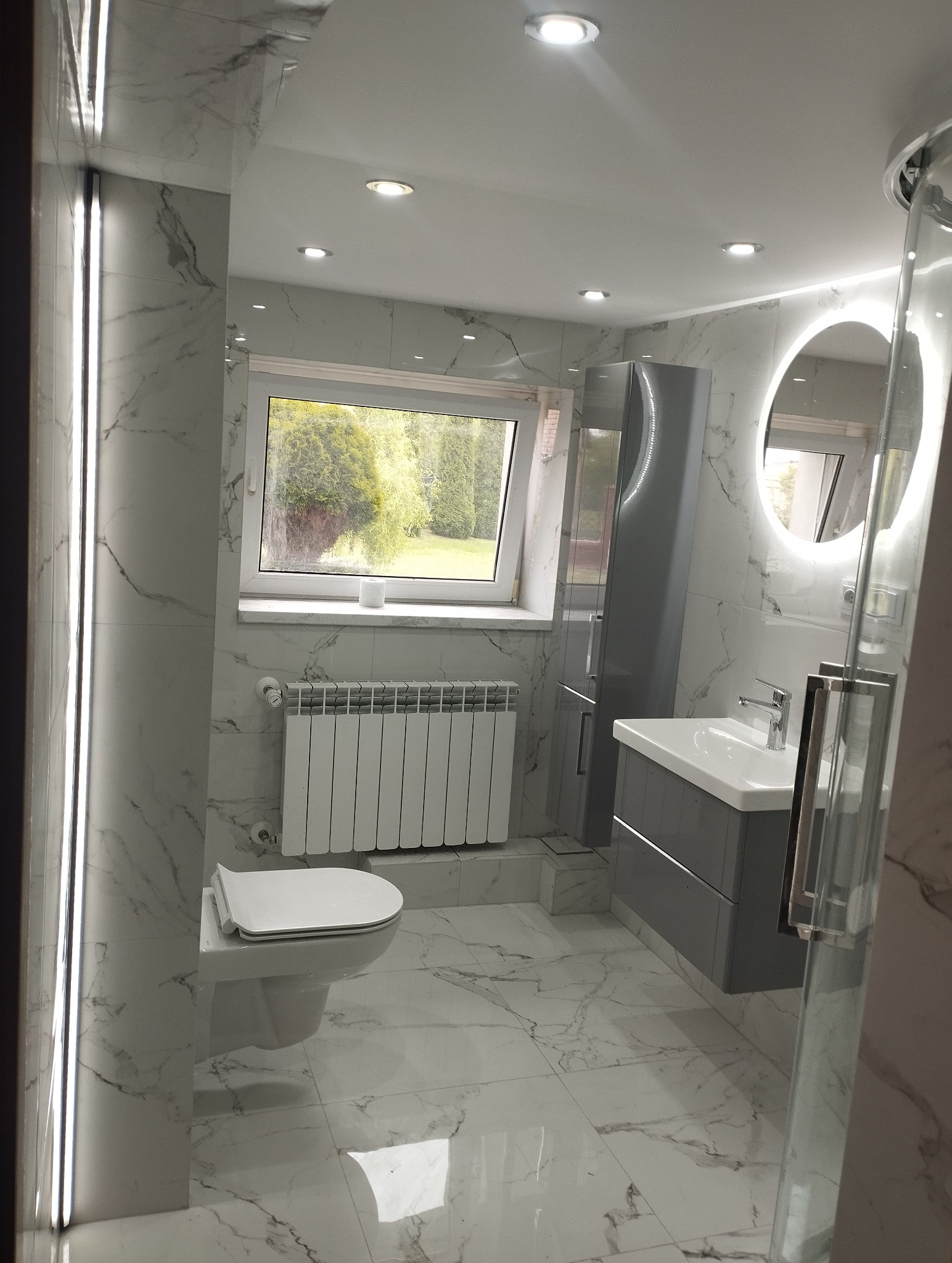 Nowoczesna łazienka z marmurowymi płytkami, szarą szafką pod umywalką i podświetlanym lustrem. Widok na okno z zielenią. Minimalistyczny design.