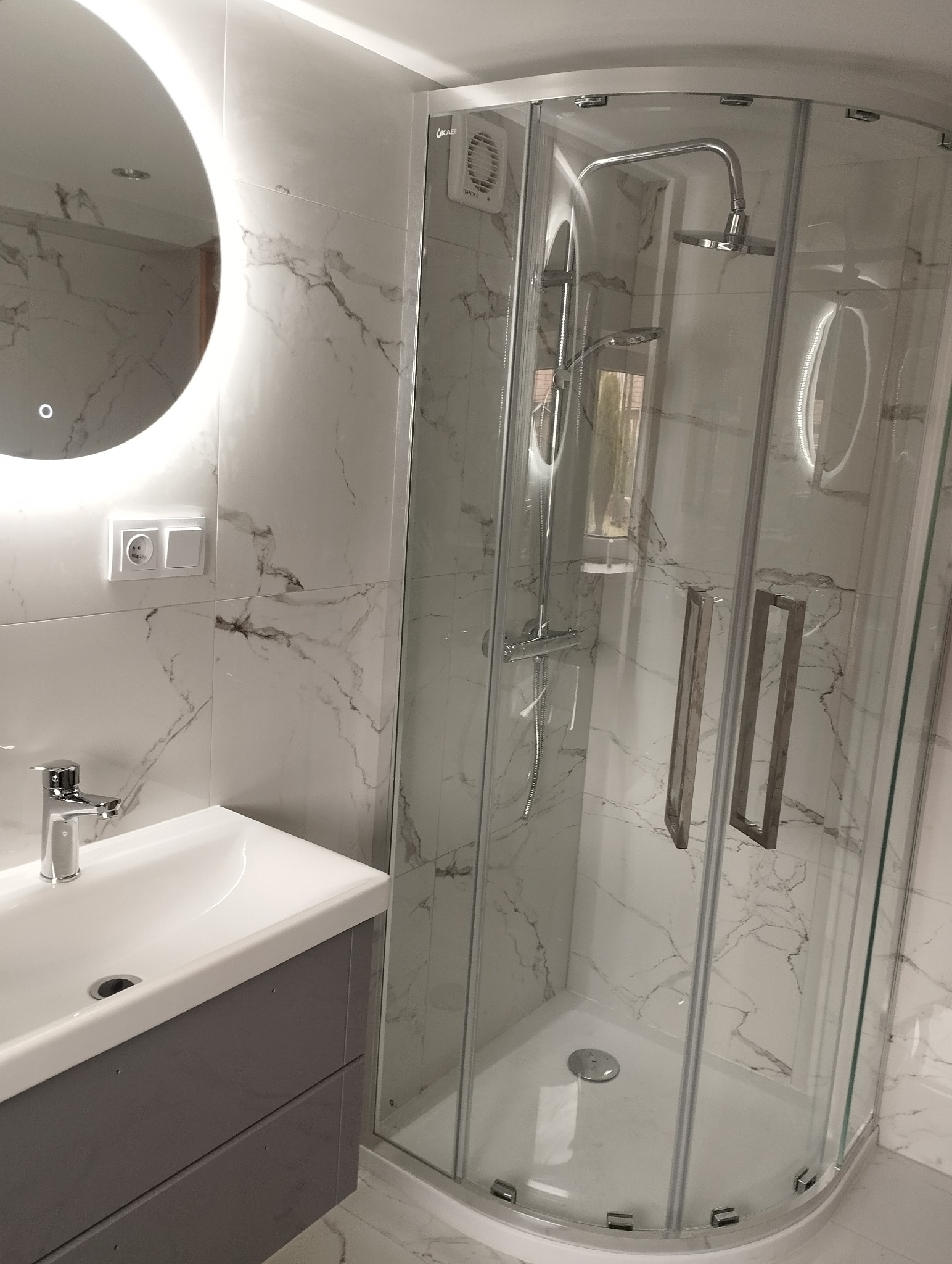 Nowoczesna łazienka z kabiną prysznicową narożną, umywalką z szafką w odcieniach szarości i okrągłym lustrem z podświetleniem LED. Marmurowe płytki na ścianach.