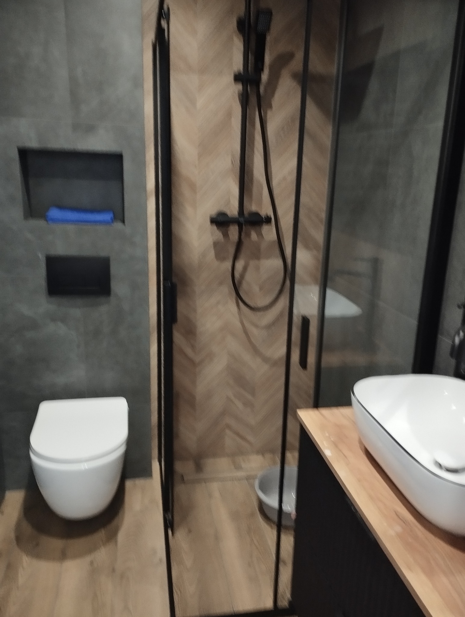 Nowoczesna łazienka z kabiną prysznicową typu walk-in, toaletą i umywalką nablatową. Ściany wykończone płytkami w odcieniach szarości i drewna.