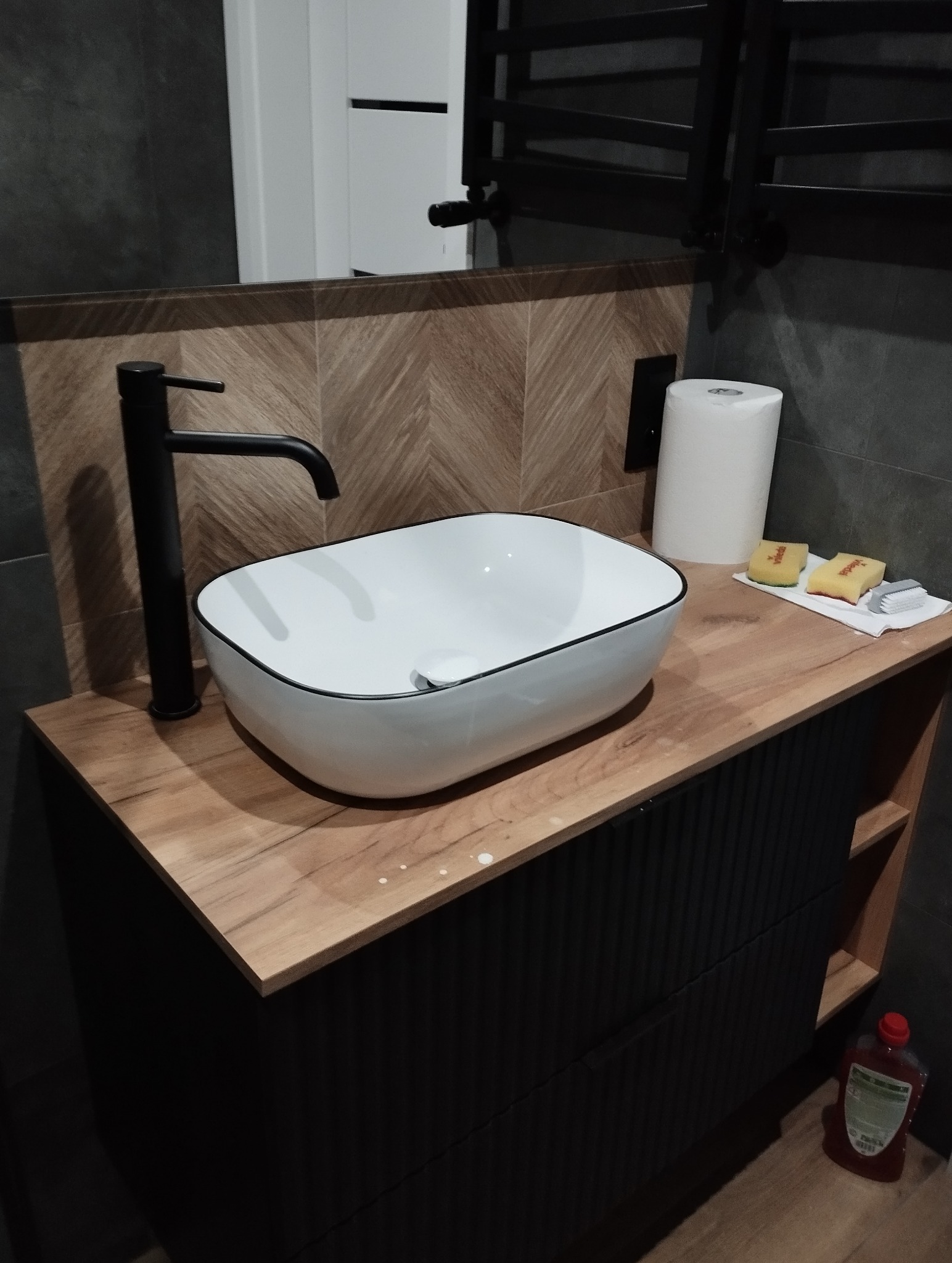 Nowoczesna łazienka: biała umywalka nablatowa z czarną armaturą na drewnianym blacie, wzorzyste płytki na ścianie i czarne szafki. Widoczne akcesoria do czyszczenia.