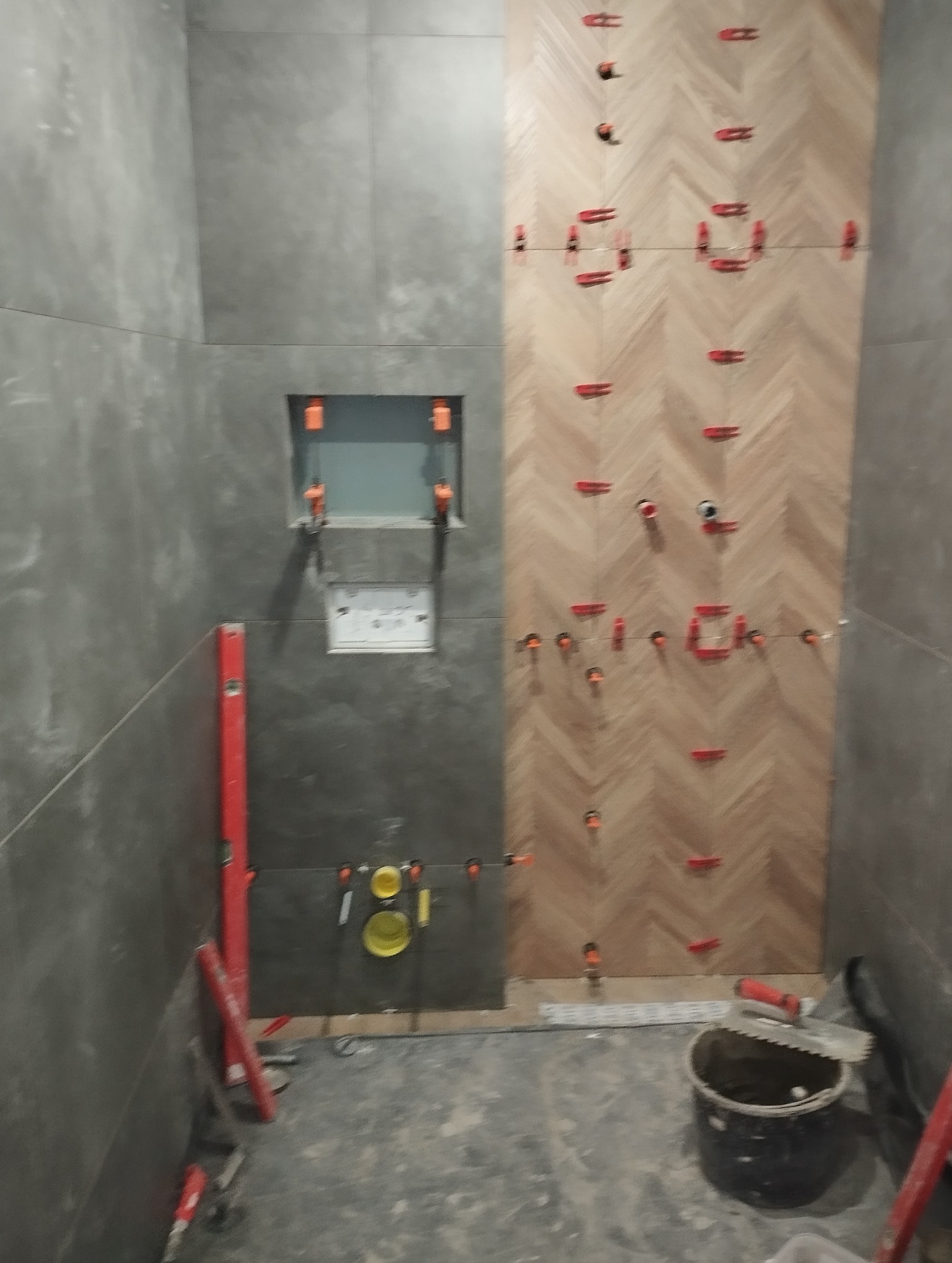 Układanie płytek w jodełkę na ścianie łazienki, z użyciem klipsów poziomujących. Część ściany wykończona szarymi płytkami. Widoczne narzędzia glazurnicze.