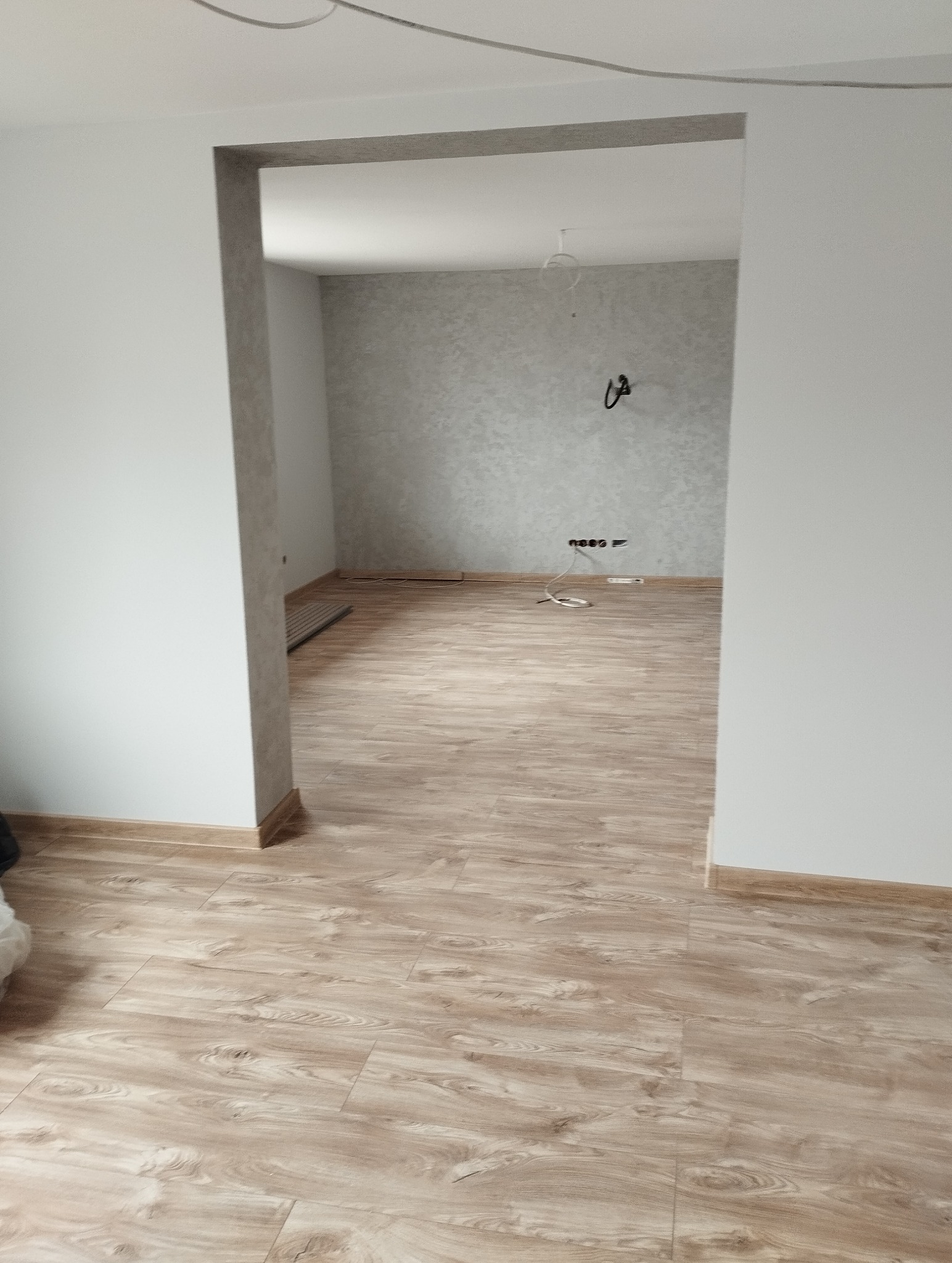 Wykończone wnętrze pokoju z panelami podłogowymi w odcieniach brązu i beżu. Ściany pomalowane na biało, jedna ściana z szarą tapetą. Widoczne gniazdka elektryczne.