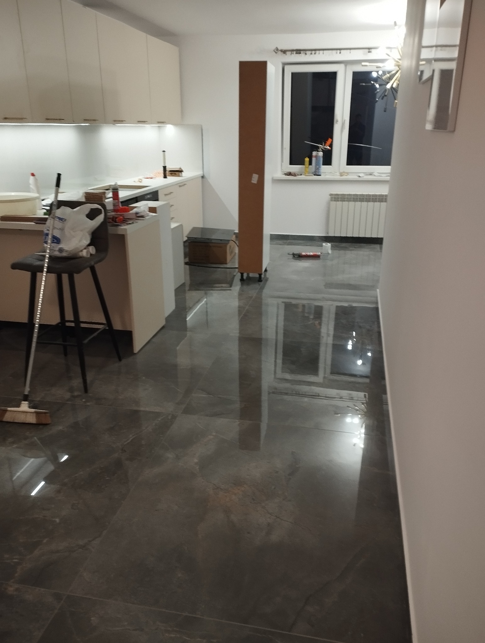 Położone, błyszczące, grafitowe płytki podłogowe w kuchni. Widoczny fragment mebli kuchennych, okno i karton. Na podłodze leży mop.