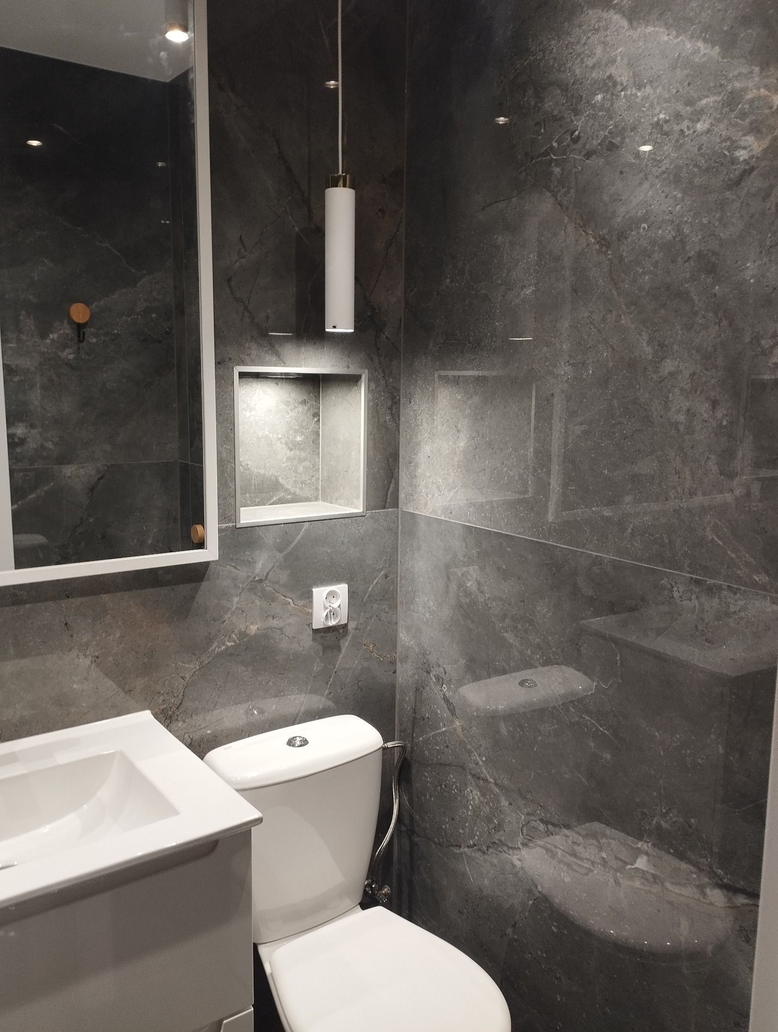 Nowoczesna łazienka z szarymi płytkami imitującymi marmur, wiszącą lampą tubą i wnęką z oświetleniem. Biała umywalka z szafką i toaleta.