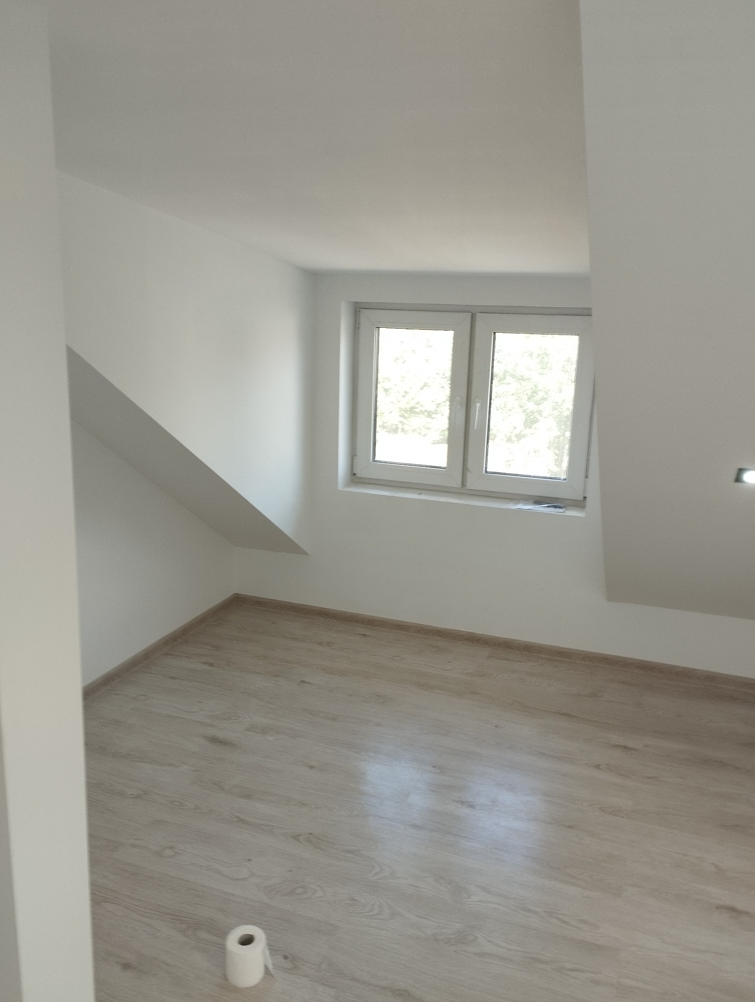 Jasne wnętrze poddasza z oknem, świeżo po remoncie. Parkiet w odcieniach beżu, białe ściany i sufit. Na podłodze leży rolka papieru toaletowego.
