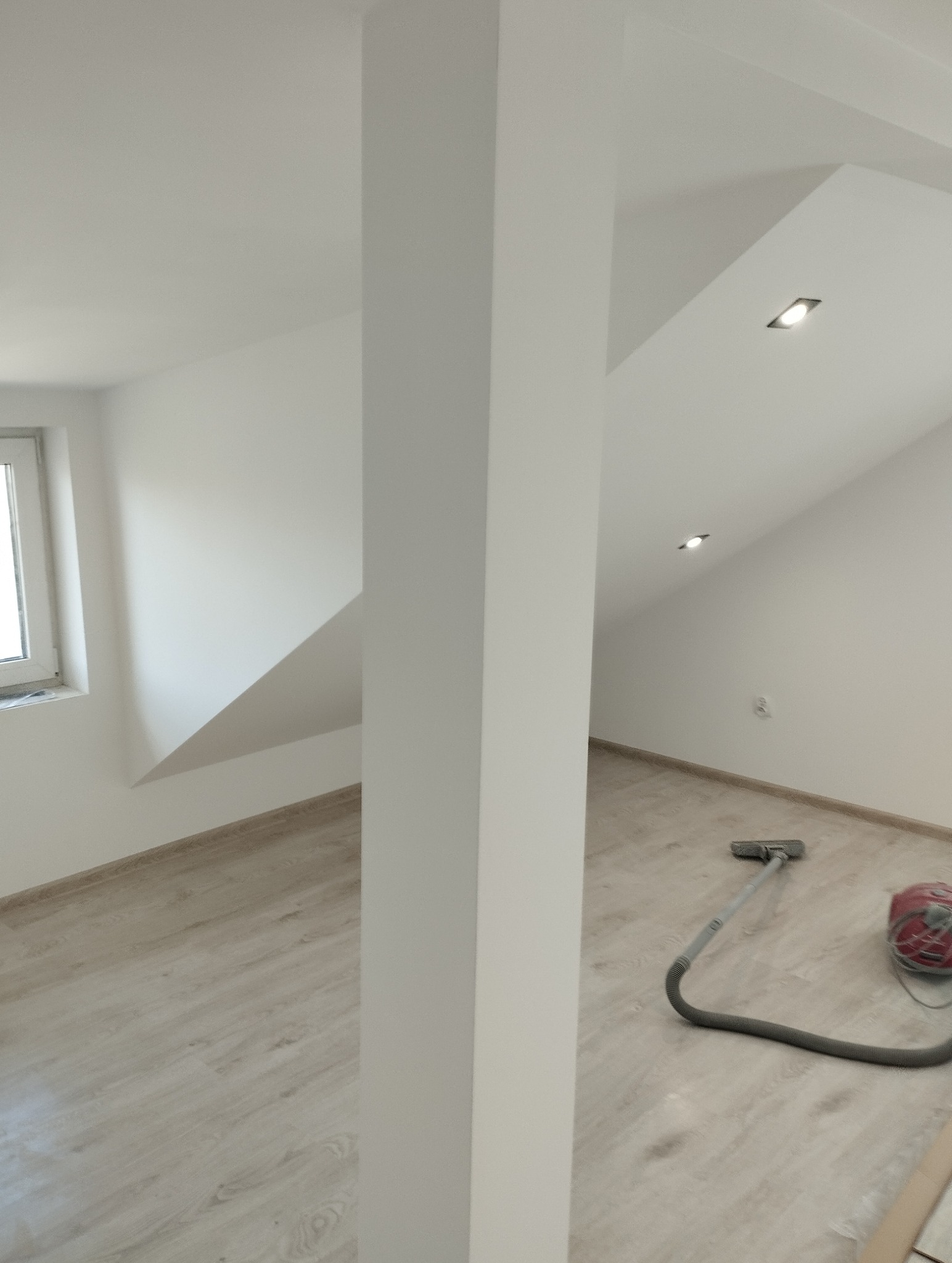 Jasne wnętrze poddasza po remoncie z drewnopodobną podłogą, kolumną i oknem. Widoczny odkurzacz. Minimalistyczne oświetlenie punktowe.