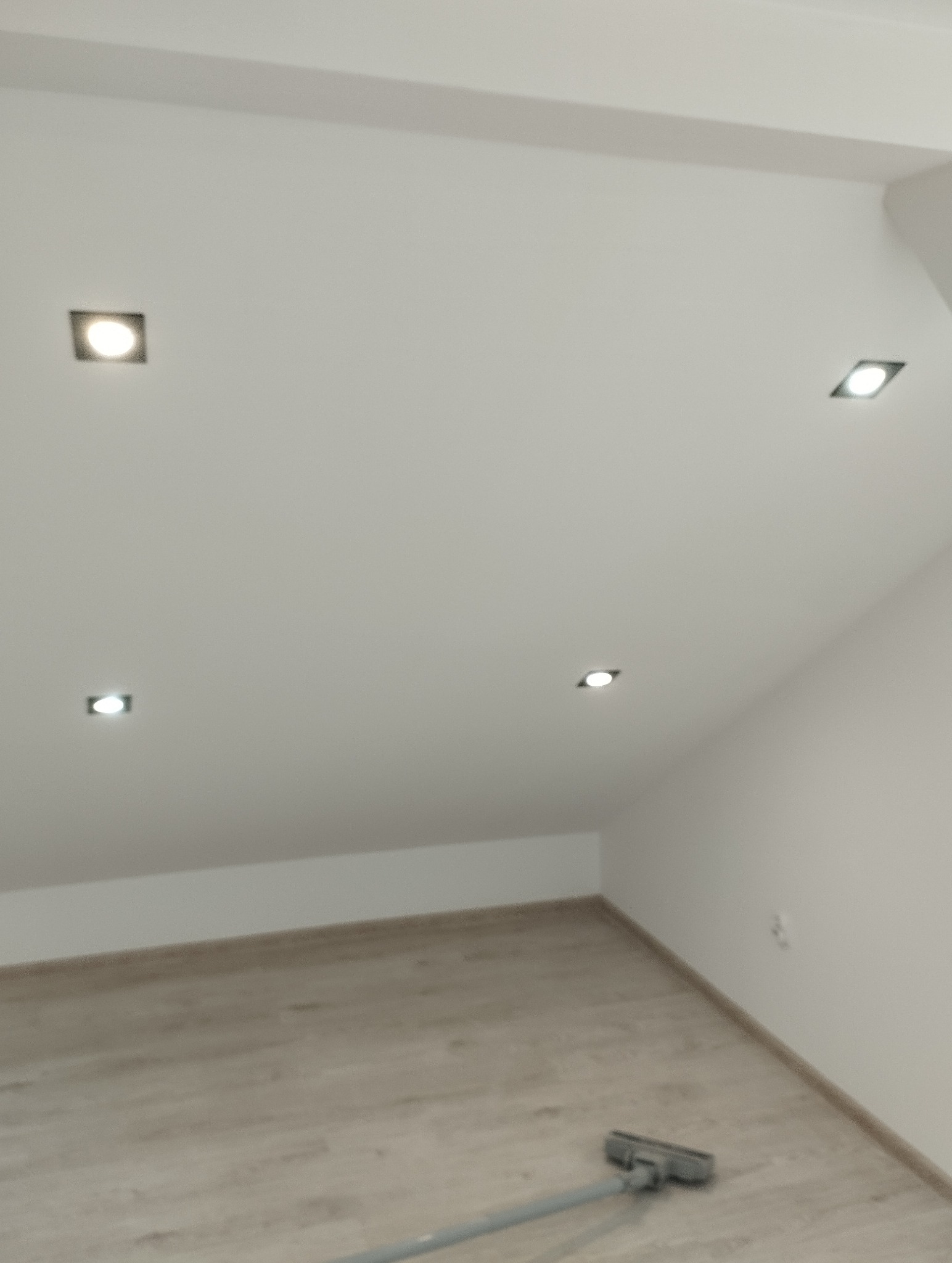 Biały, skośny sufit poddasza z czterema kwadratowymi lampami LED. Na podłodze widoczny odkurzacz. Minimalistyczne wykończenie wnętrza.