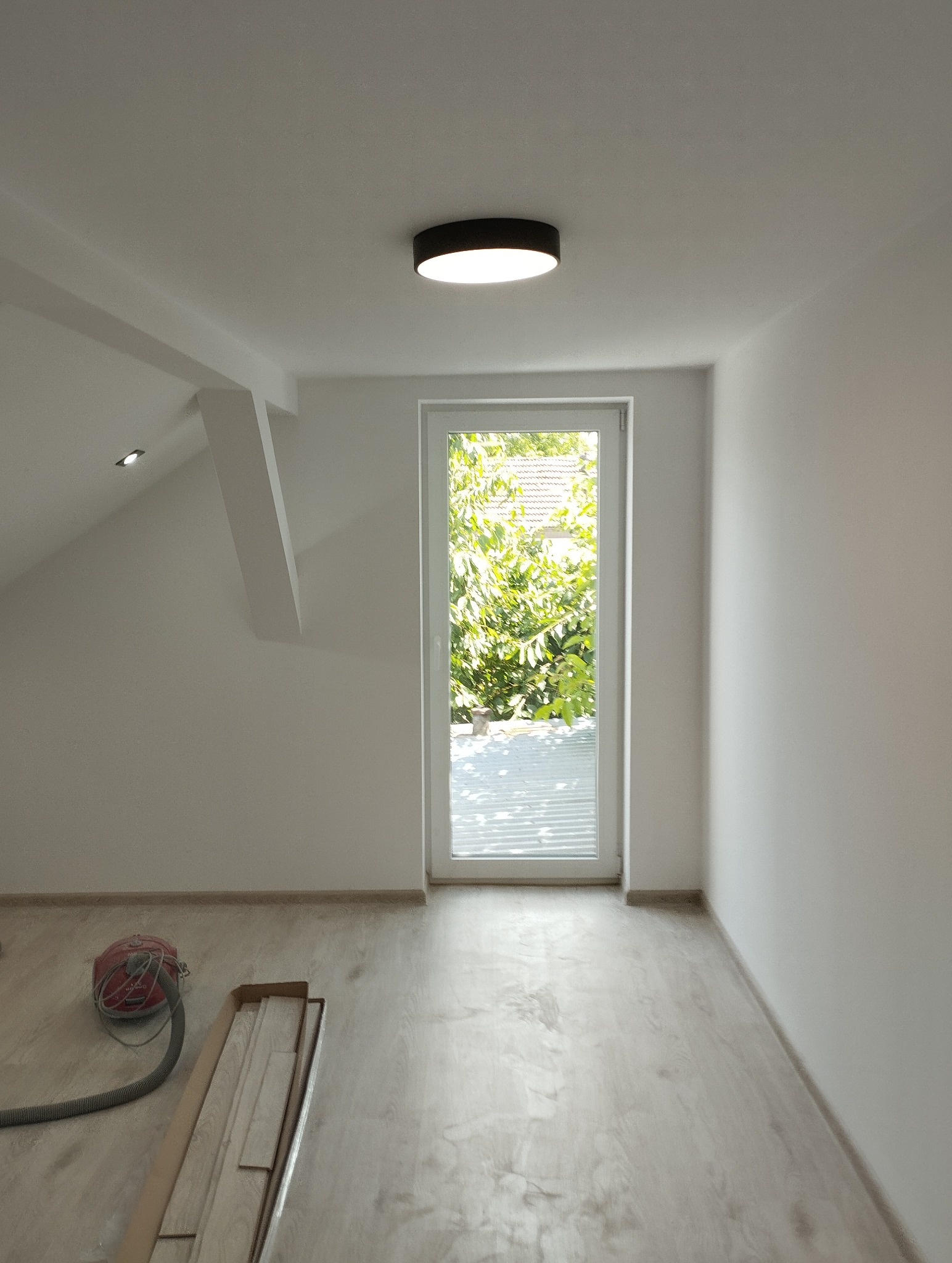 Jasne poddasze z oknem i nową podłogą. Widoczny odkurzacz i panele podłogowe w kartonie. Minimalistyczna lampa sufitowa. Świeżo wykończone wnętrze.