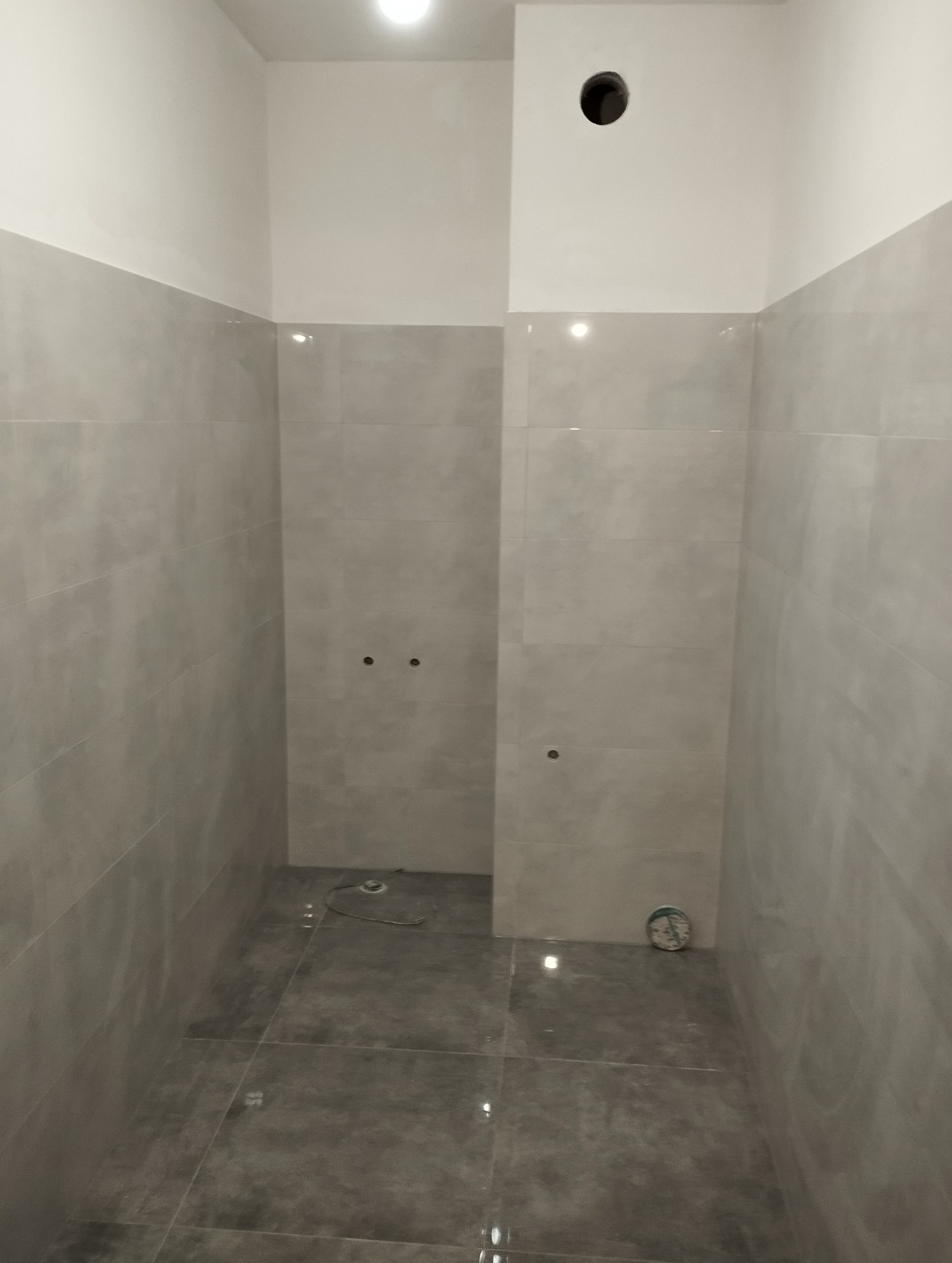 Wnętrze łazienki w trakcie prac wykończeniowych: szare płytki na ścianach i podłodze, widoczne otwory na armaturę, sufit pomalowany na biało.