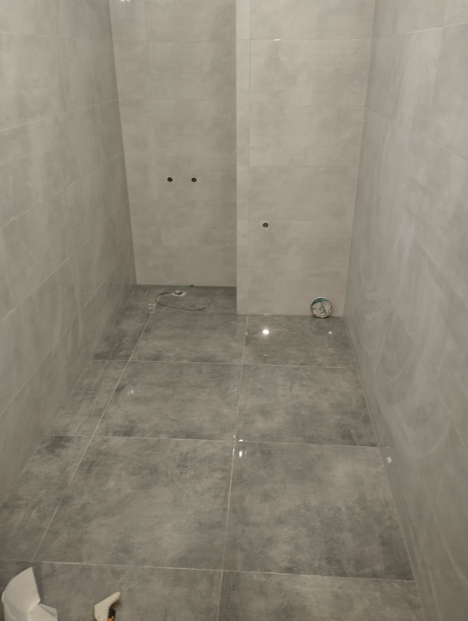 Wykończona łazienka z szarymi, matowymi płytkami na ścianach i podłodze. Widoczne otwory na armaturę. Minimalistyczny design, świeżo po remoncie.