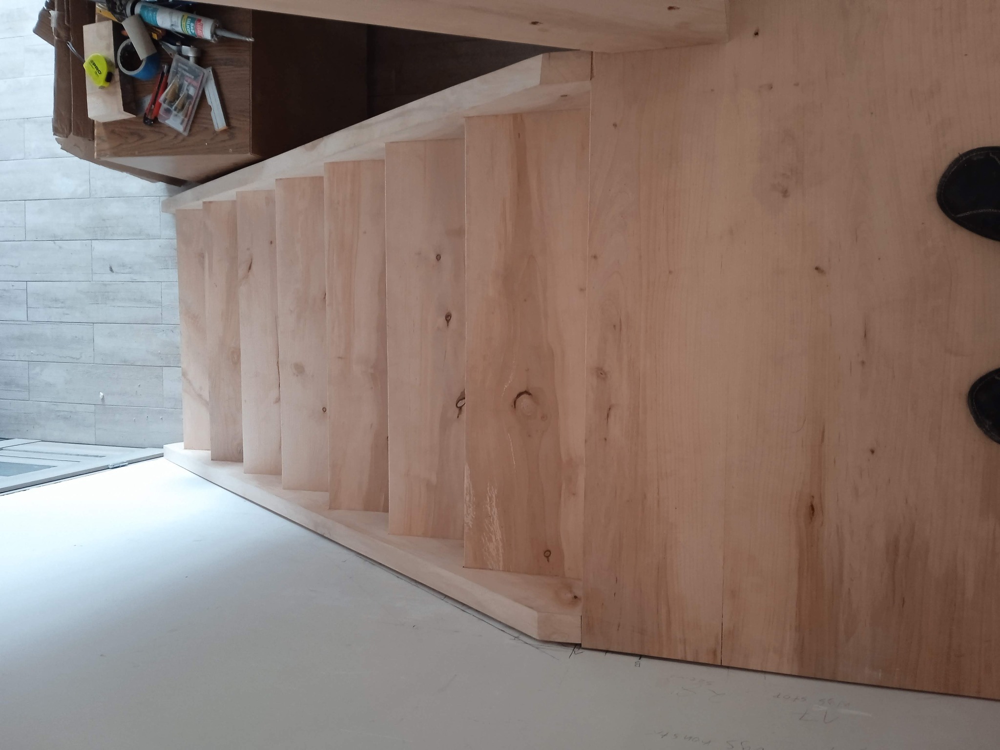 Drewniana konstrukcja schodów w trakcie montażu, widoczne narzędzia i materiały. Jasne drewno, minimalistyczne wnętrze, częściowo widoczne stopy osoby.