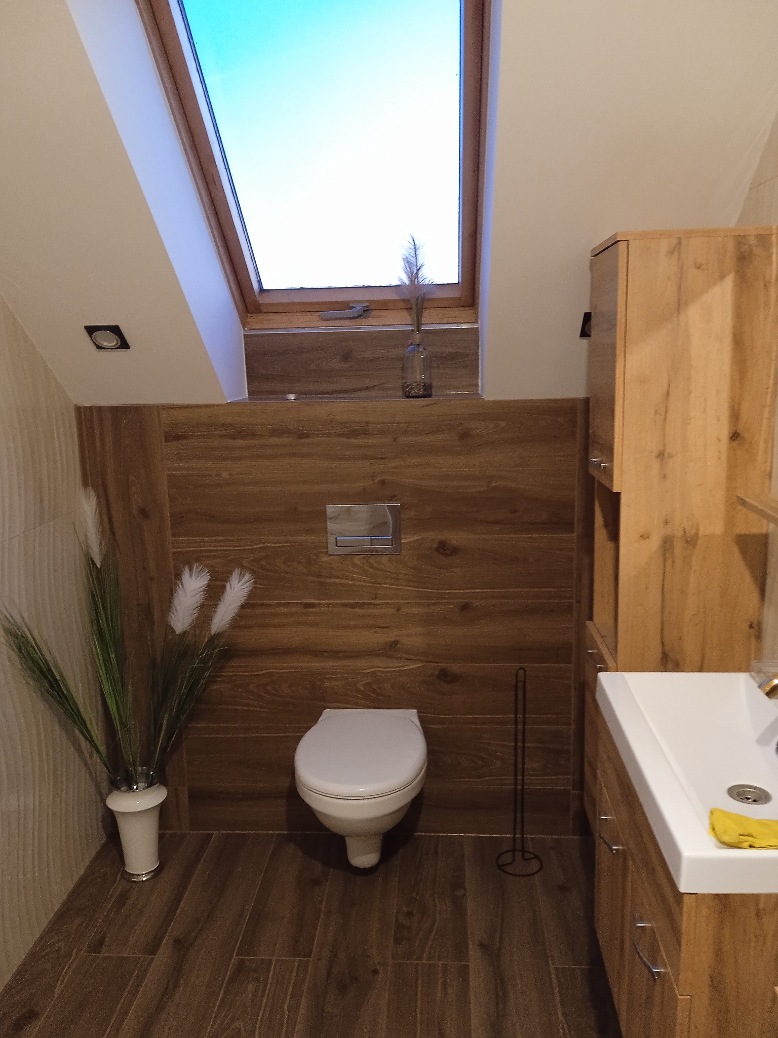 Drewnopodobna łazienka na poddaszu z oknem dachowym. Umywalka z szafką, toaleta, dekoracje z traw, brązowe płytki imitujące drewno na podłodze i ścianie.