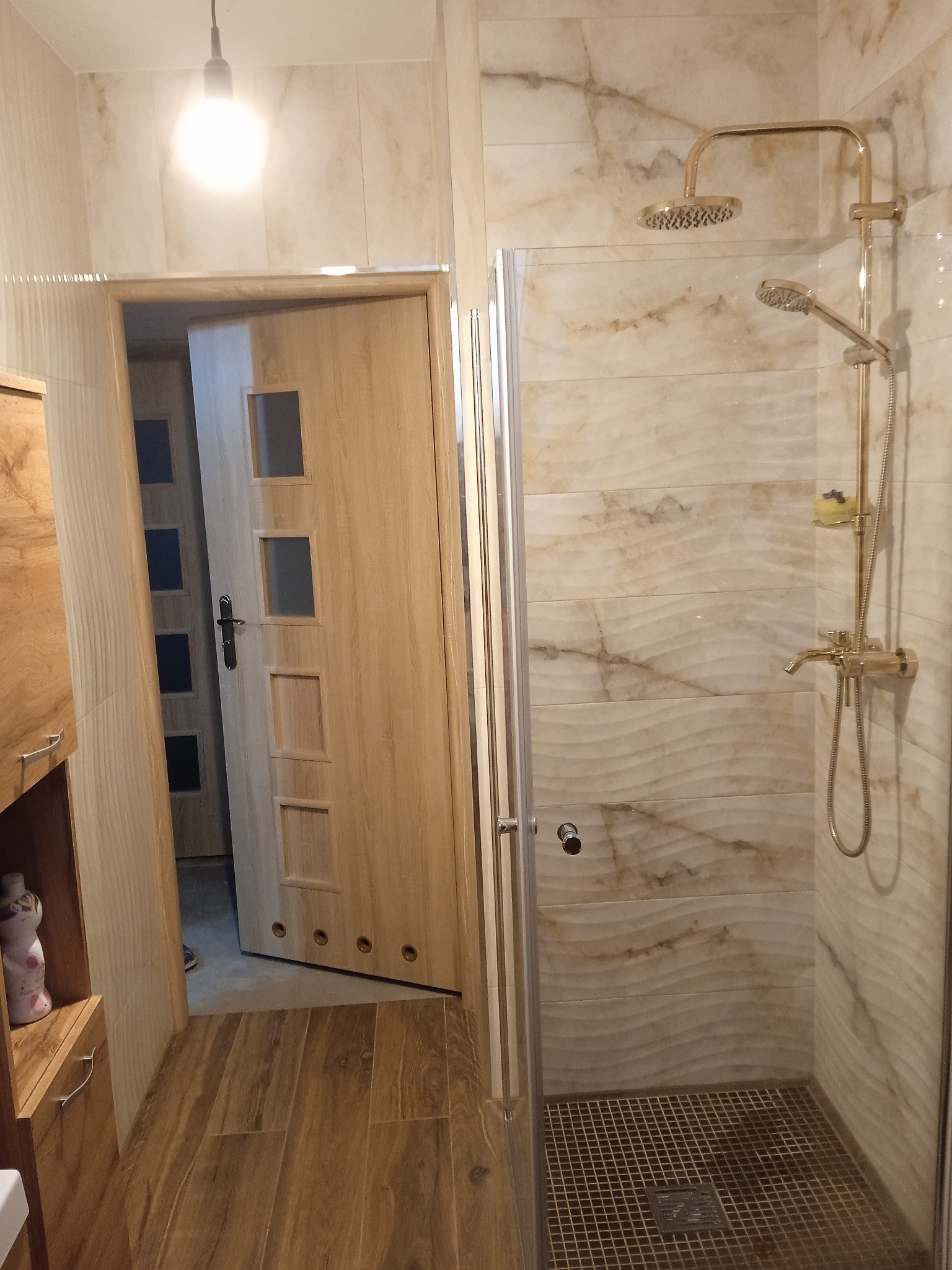 Wykończona łazienka z kabiną prysznicową, złotą armaturą i beżowymi płytkami imitującymi marmur. Widoczne drewniane drzwi z przeszkleniami i szafka.