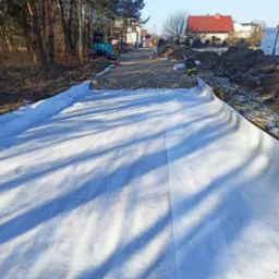 Przygotowanie podłoża pod drogę dojazdową: rozłożona geowłóknina na podmokłym terenie, w tle widoczny mały koparka i budynki mieszkalne.