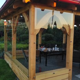 Zakład Szklarski Tęcza - Zadaszona altana ogrodowa z drewnianą konstrukcją i szklanymi panelami bocznymi mocowanymi za pomocą metalowych śrub, widoczne odbicie drzew w szkle, na stole wewnątrz altany naczynia i butelka...