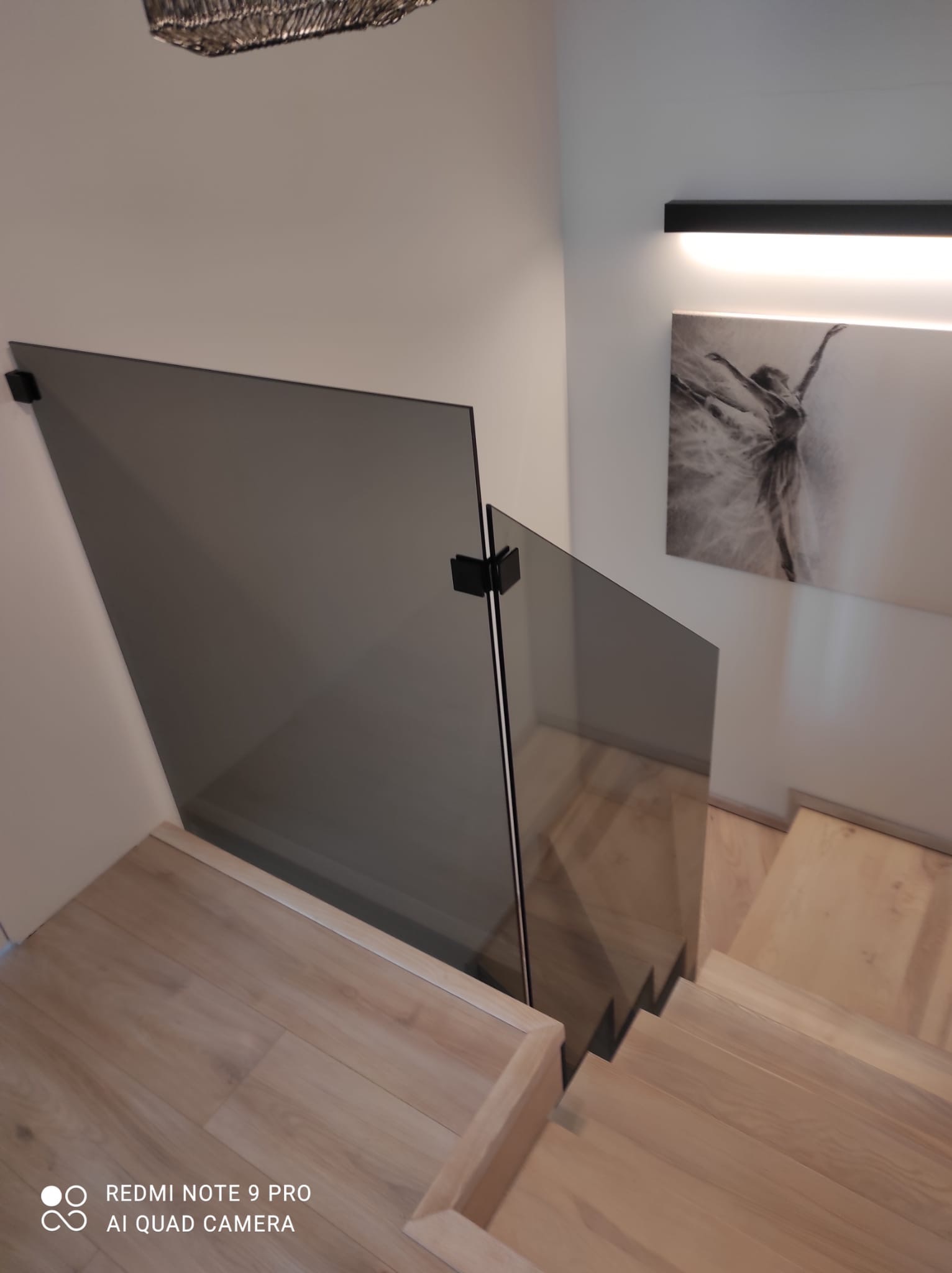 Szara balustrada szklana na drewnianych schodach w nowoczesnym wnętrzu, z czarnymi uchwytami i obrazem baletnicy na ścianie.