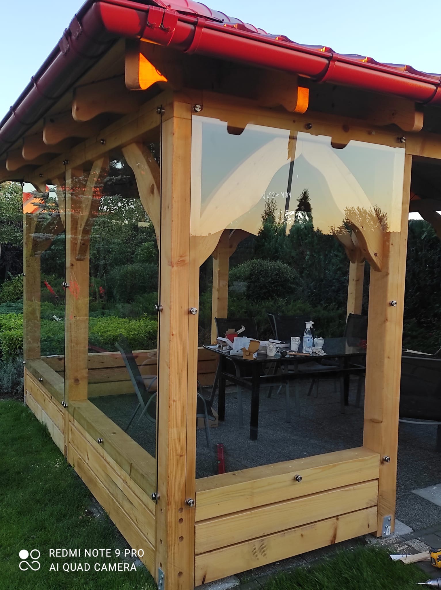 Zadaszona altana ogrodowa z drewnianą konstrukcją i szklanymi panelami bocznymi mocowanymi za pomocą metalowych śrub, widoczne odbicie drzew w szkle, na stole wewnątrz altany naczynia i butelka...