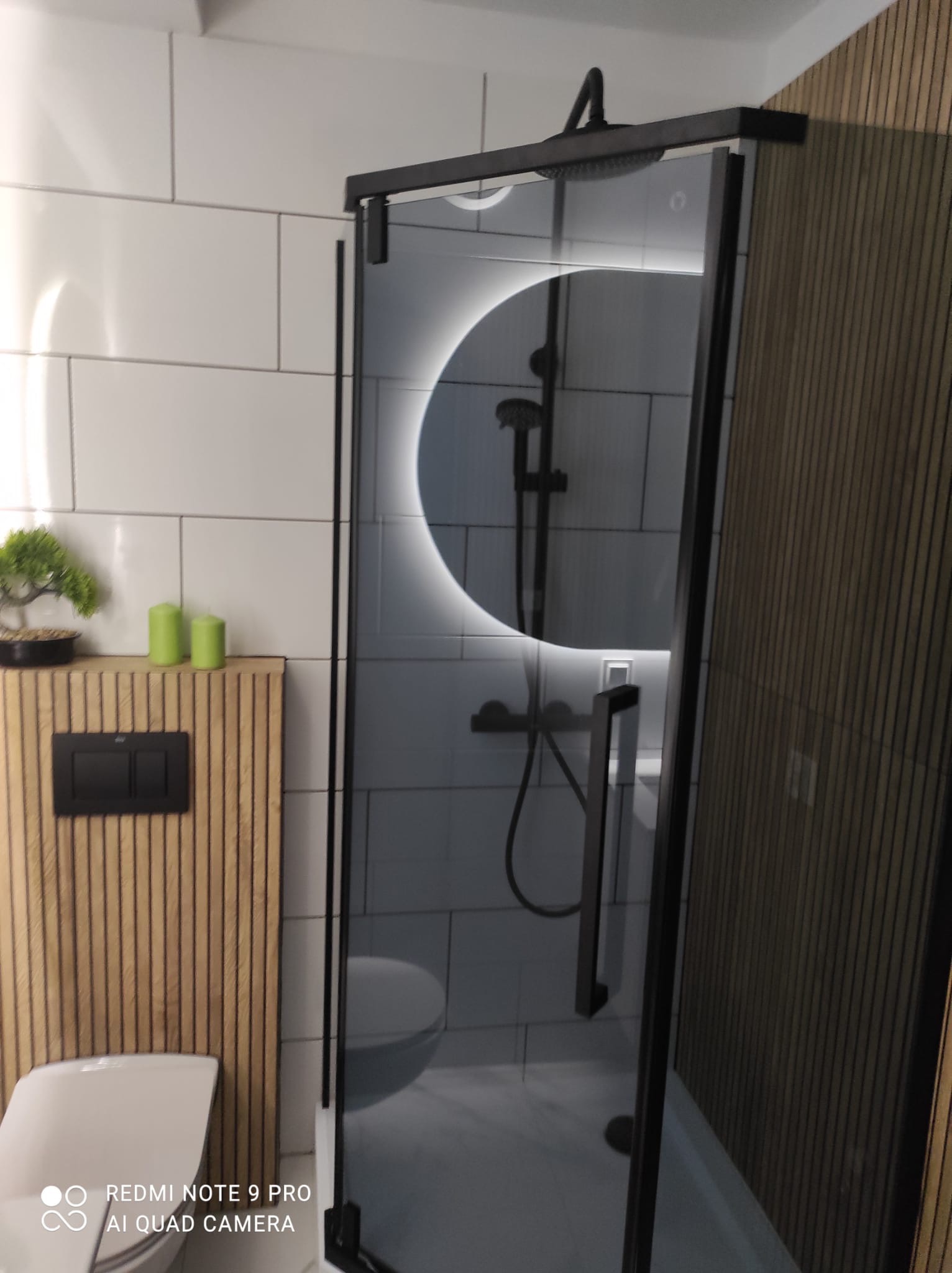 Nowoczesna kabina prysznicowa z czarną ramą i okrągłym lustrem z podświetleniem LED, widok na toaletę i drewniane panele ścienne, akcenty zieleni w postaci świec i bonsai