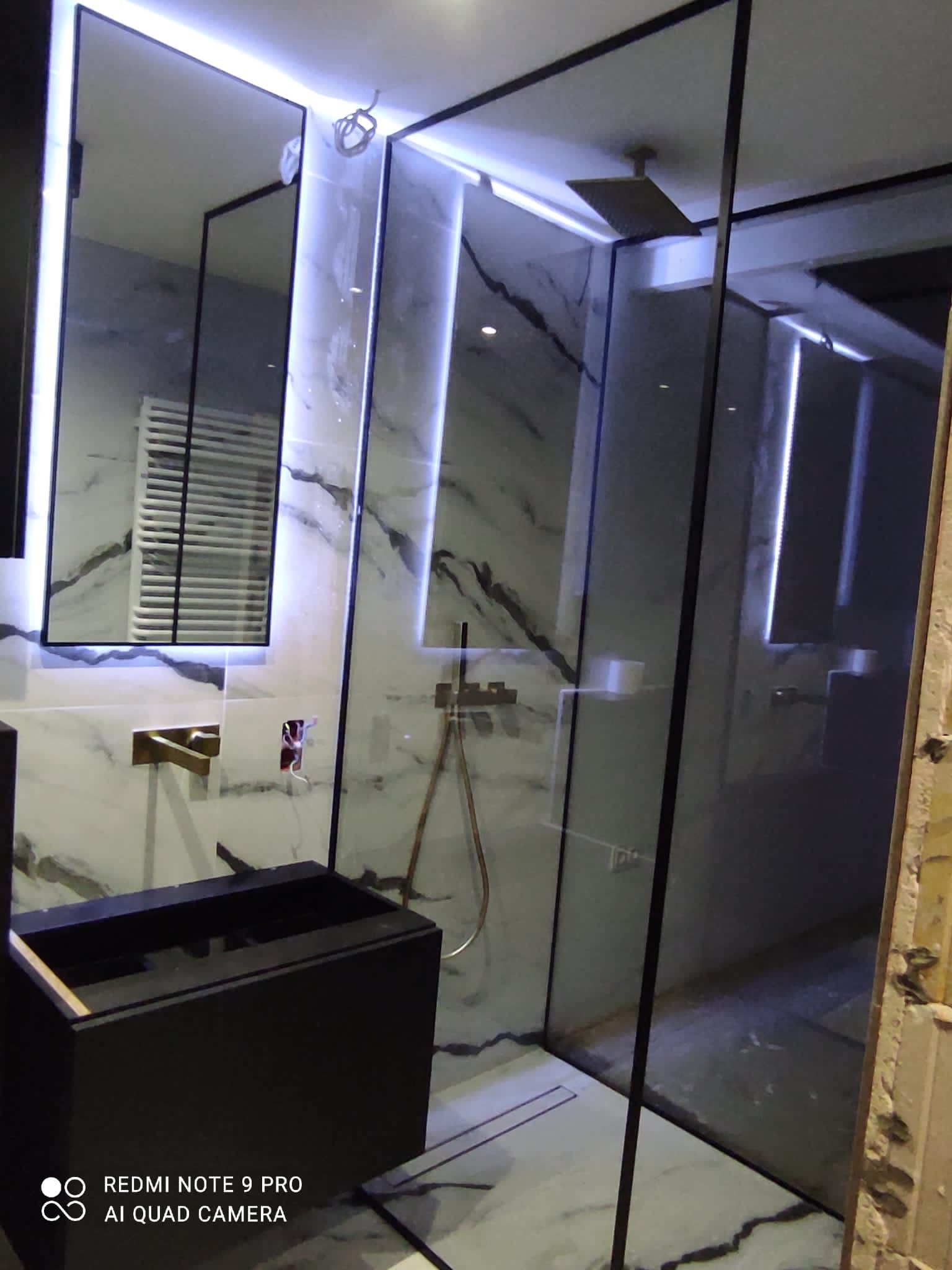 Nowoczesna łazienka z kabiną prysznicową ze szkła hartowanego z czarnymi ramami, marmurowymi płytkami i podświetleniem LED. Widoczna umywalka z czarnym blatem i bateria w kolorze szczotkowanego...