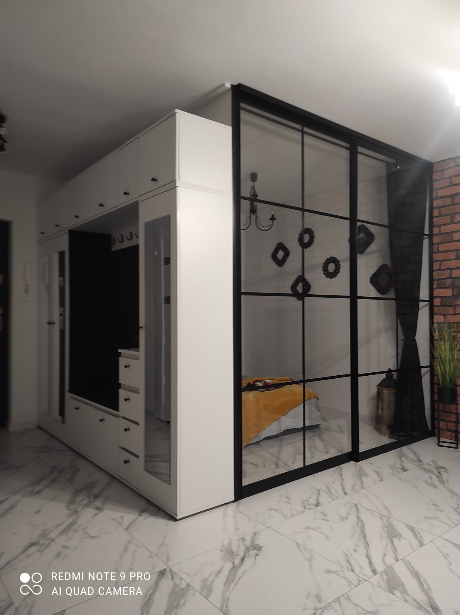 Biała szafa wnękowa z lustrami i przeszklona ścianka działowa z czarną ramą, oddzielająca sypialnię, widoczna ceglana ściana w tle.