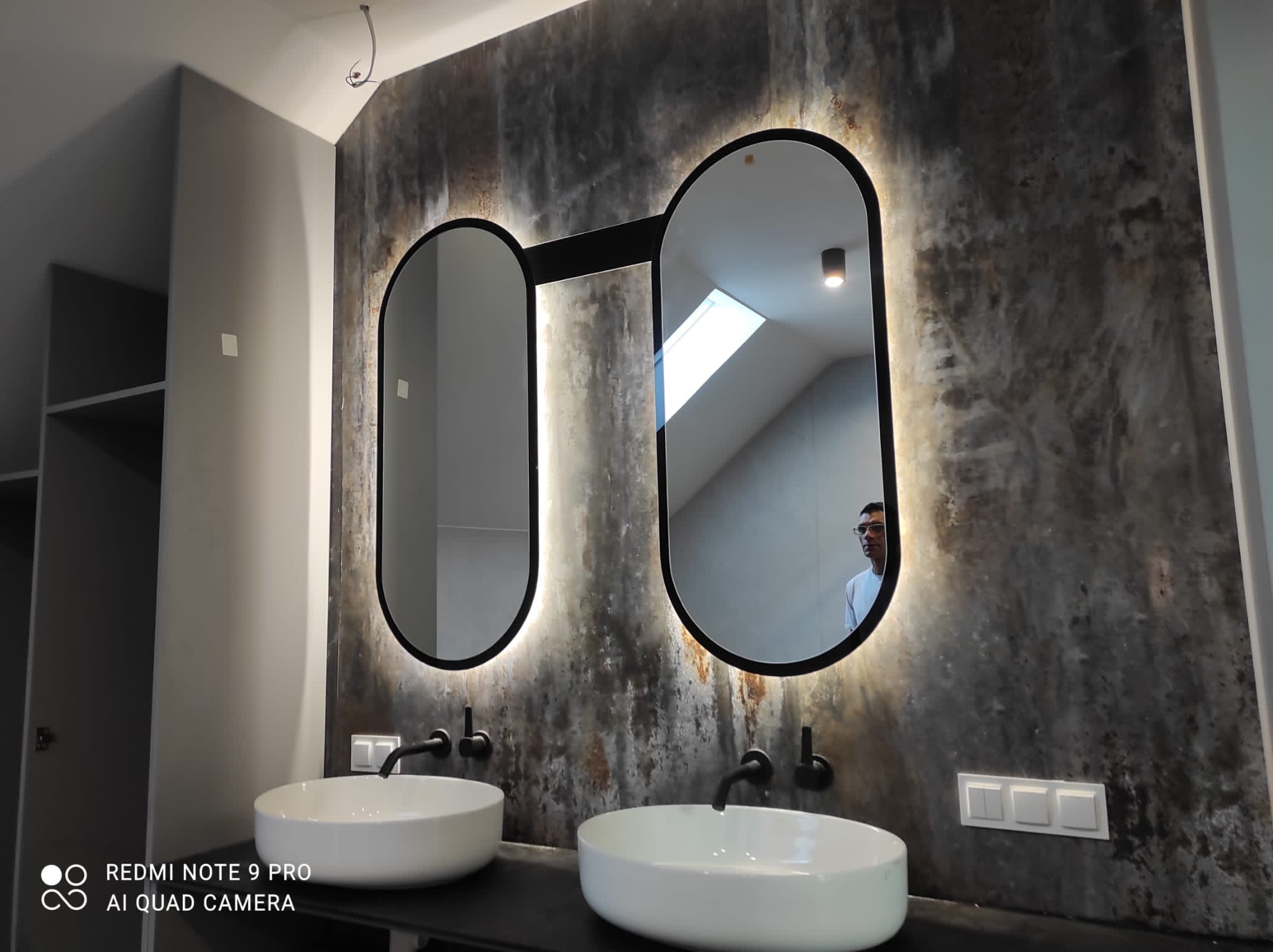 Dwie umywalki nablatowe z czarnymi bateriami i podświetlanymi lustrami w kształcie zaokrąglonych prostokątów na ścianie z efektem betonu. W lustrze odbija się osoba.