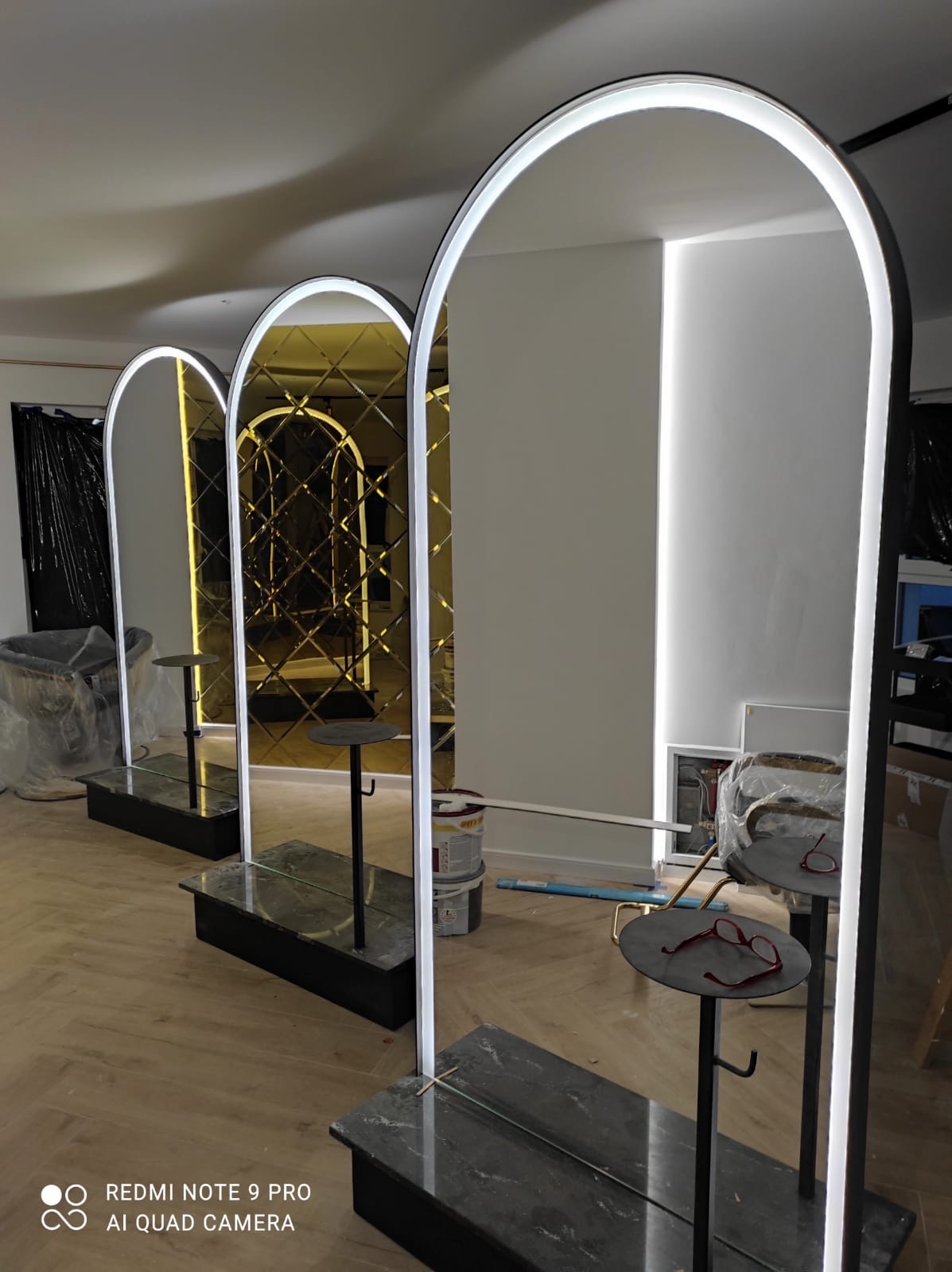 Trzy podświetlane lustra łukowe z podstawami z czarnego marmuru, ustawione w rzędzie w pomieszczeniu w trakcie wykańczania, jedno z nich z dekoracyjną, złoconą powierzchnią lustrzaną, na jednej...