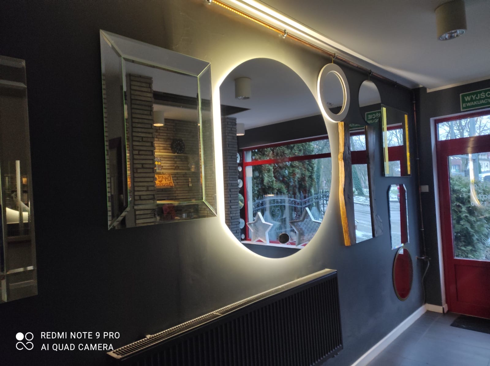 Różne lustra dekoracyjne na szarej ścianie, w tym podświetlane okrągłe lustro i prostokątne lustro z odbiciem ceglanej ściany w tle, nad grzejnikiem.