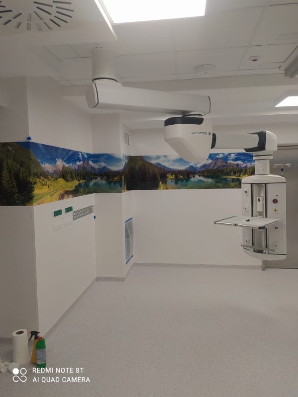 Wyposażenie sali operacyjnej: ramię sufitowe z oświetleniem i panelem sterowania, dekoracyjny pas z motywem górskiego krajobrazu, białe ściany i szara podłoga z widocznymi śladami po taśmie...