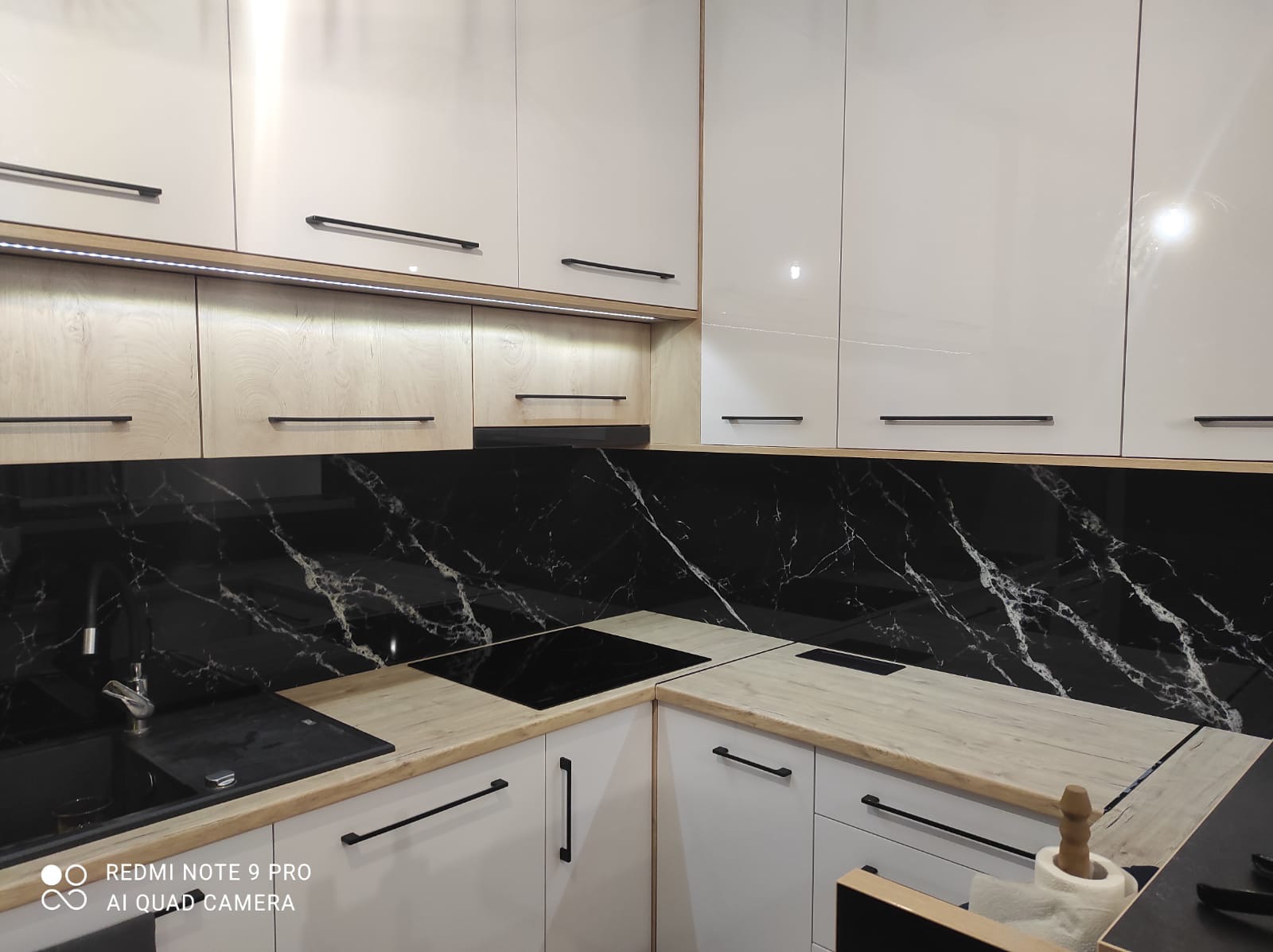 Nowoczesna kuchnia z białymi błyszczącymi szafkami, drewnianymi blatami i czarnym panelem szklanym imitującym marmur nad blatem. Oświetlenie LED pod szafkami wiszącymi.