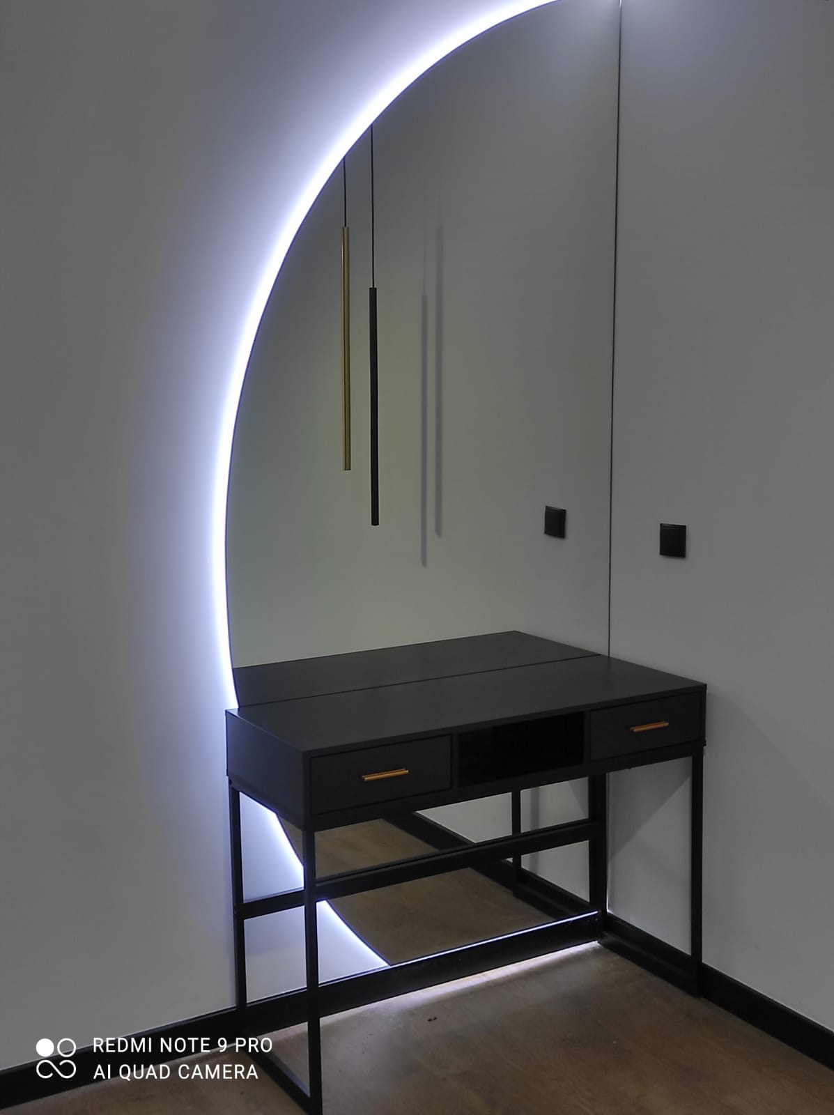 Czarna toaletka z dwoma szufladami i lustrem podświetlonym ledowym łukiem, wiszące lampy-tuby w tle, minimalistyczny design.