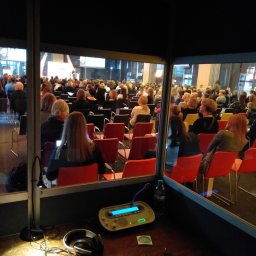 Marcelina Bochyńska-Graczyk - Widok z kabiny tłumacza symultanicznego na salę konferencyjną wypełnioną uczestnikami, na pierwszym planie słuchawki, mikrofon i konsola tłumacza.