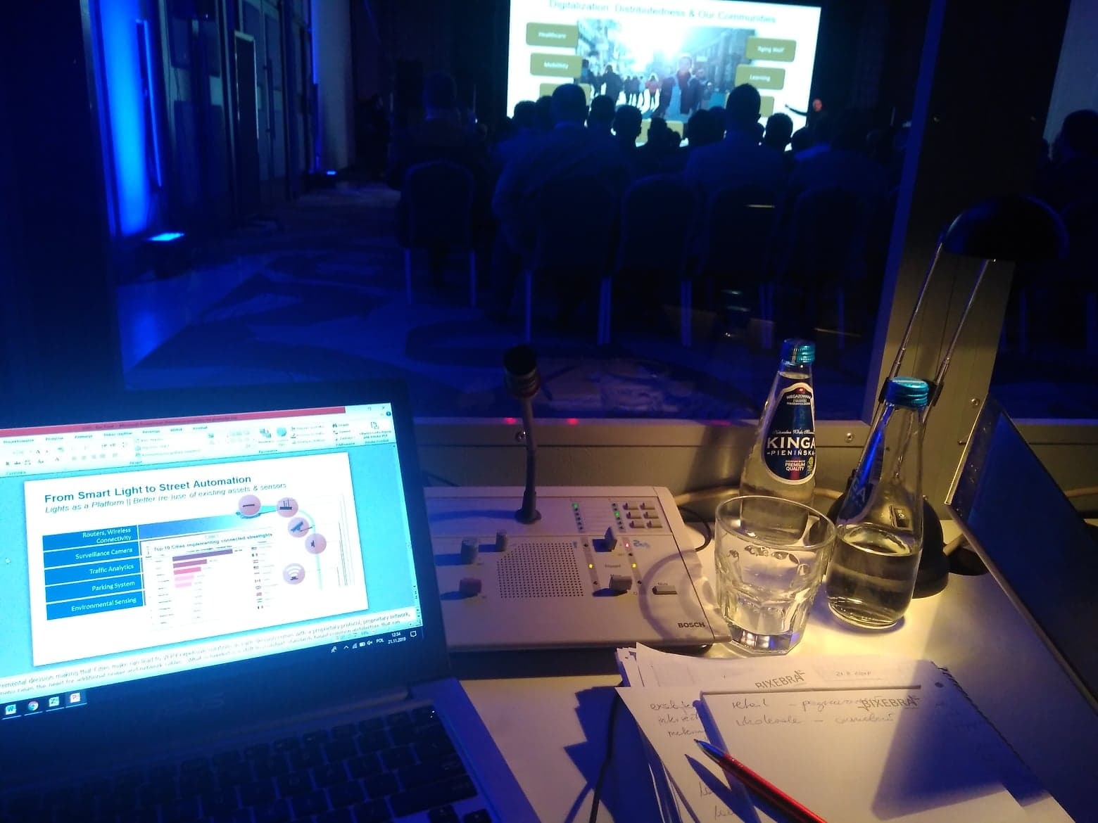 Stanowisko tłumacza konferencyjnego z laptopem wyświetlającym prezentację o inteligentnym oświetleniu ulicznym, mikrofonem, notatkami i butelkami wody na tle sali konferencyjnej z publicznością...