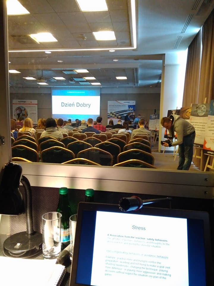 Widok zza kulis konferencji: laptop z prezentacją 'Stress' na pierwszym planie, w tle sala konferencyjna z uczestnikami i ekranem wyświetlającym 'Dzień Dobry'.