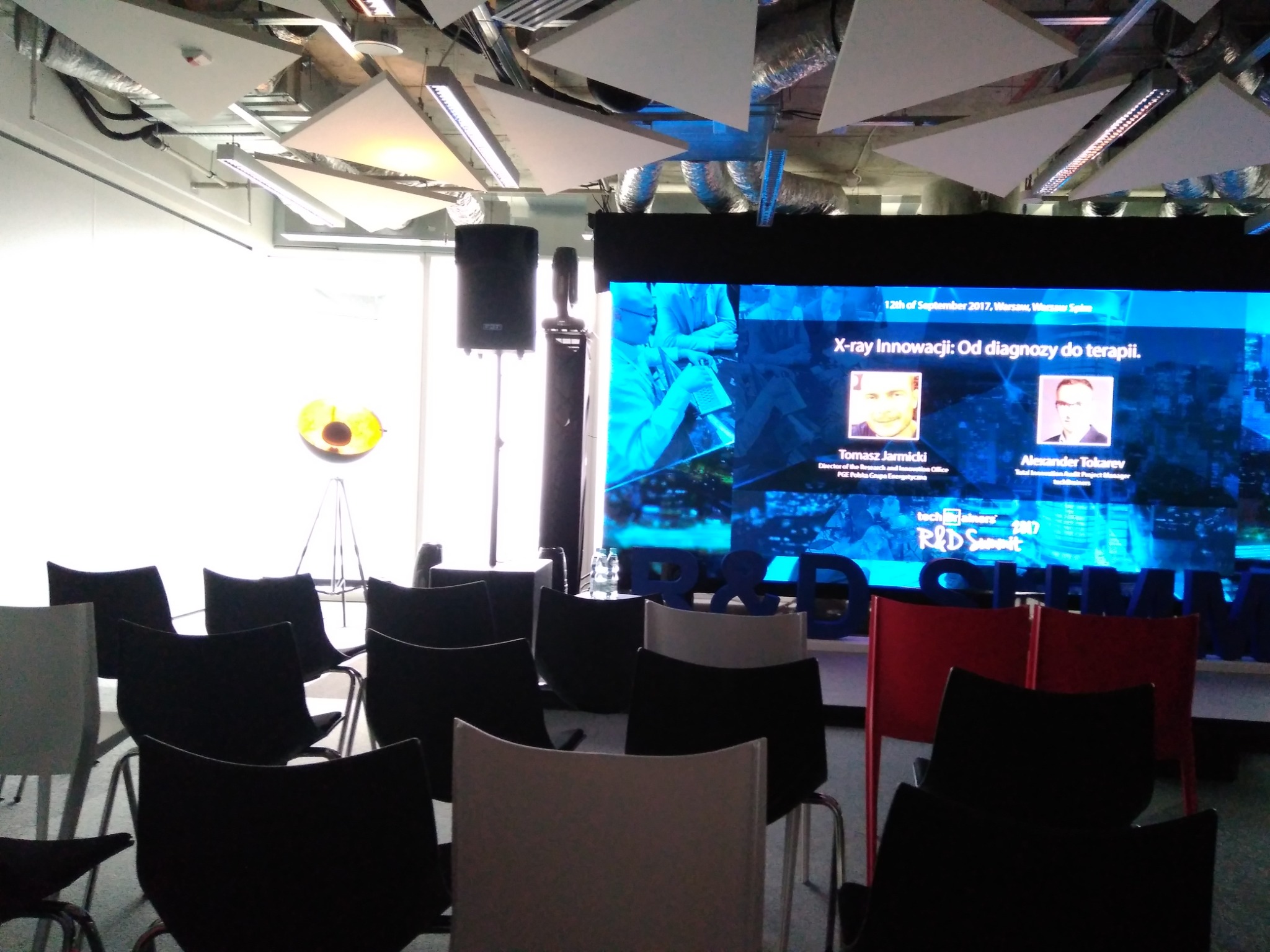 Sala konferencyjna z rzędami krzeseł przed ekranem prezentującym prelegentów i temat 'X-ray Innowacji: Od diagnozy do terapii', z elementami dekoracyjnymi na suficie i butelkami wody na stole w tle.