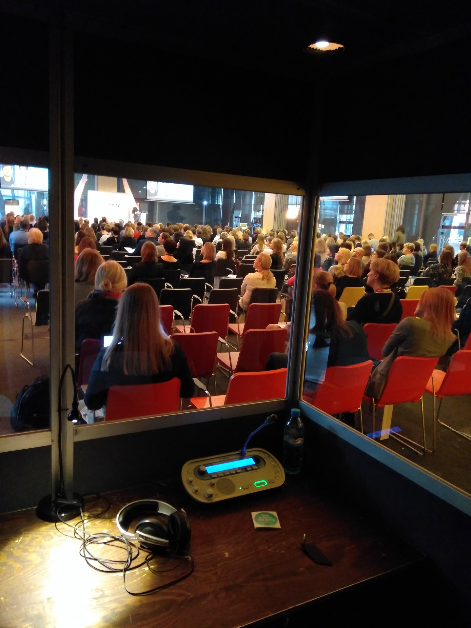 Widok z kabiny tłumacza symultanicznego na salę konferencyjną wypełnioną uczestnikami, na pierwszym planie słuchawki, mikrofon i konsola tłumacza.
