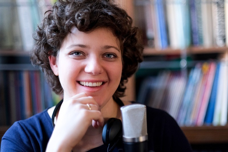 Uśmiechnięta kobieta z kręconymi włosami, mikrofonem i słuchawkami na szyi, w tle regał z książkami.