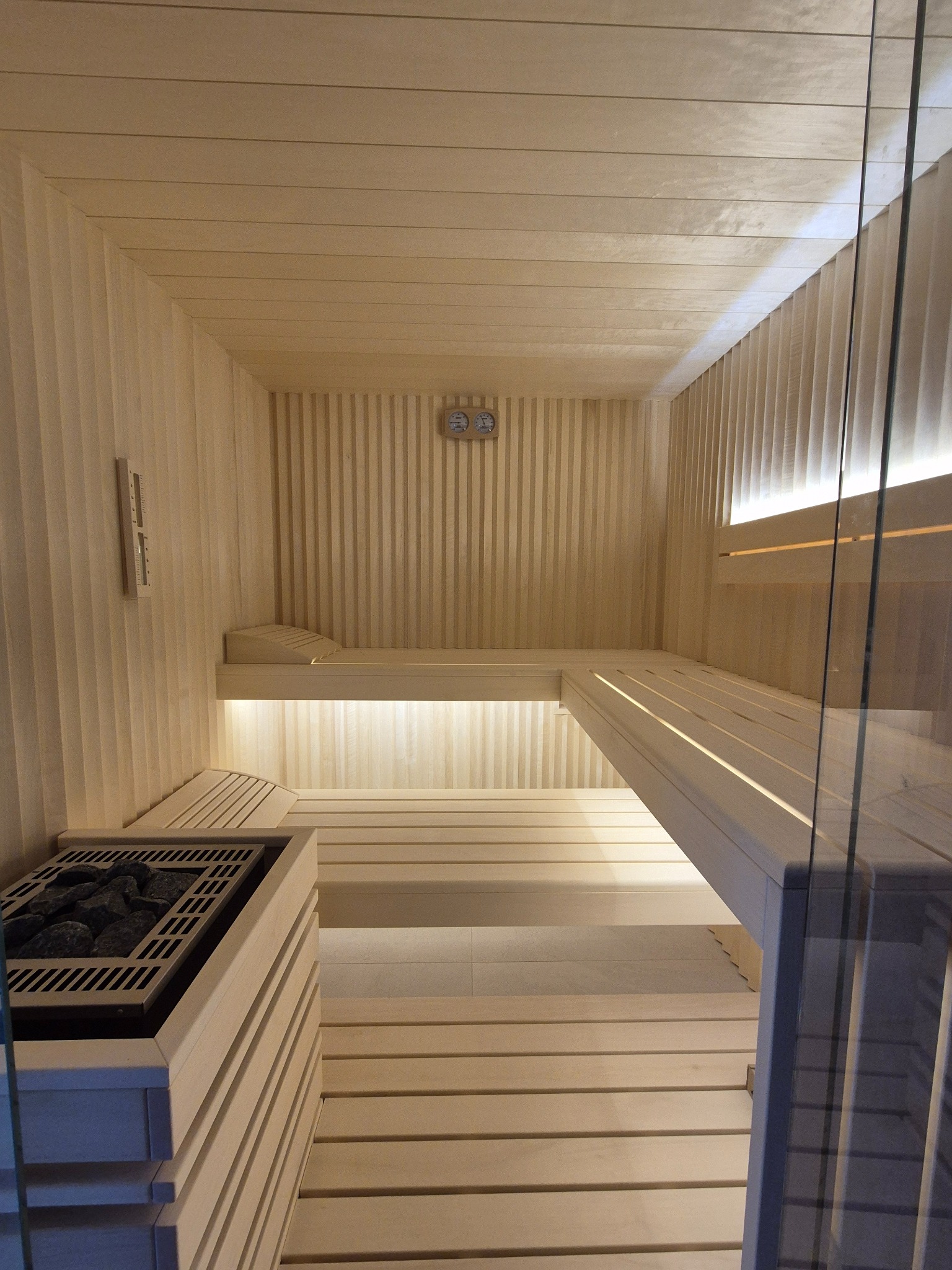 Jasne wnętrze sauny wykończone drewnem, z piecem na kamienie, termometrem i higrometrem na ścianie oraz szklaną ścianą z boku.