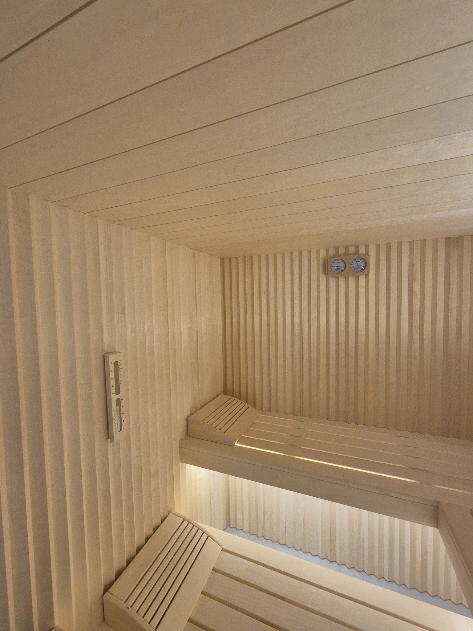 Wnętrze sauny wykończone jasnym drewnem, z dwupoziomowymi ławkami, termometrem i higrometrem na ścianie oraz subtelnym oświetleniem pod ławkami.