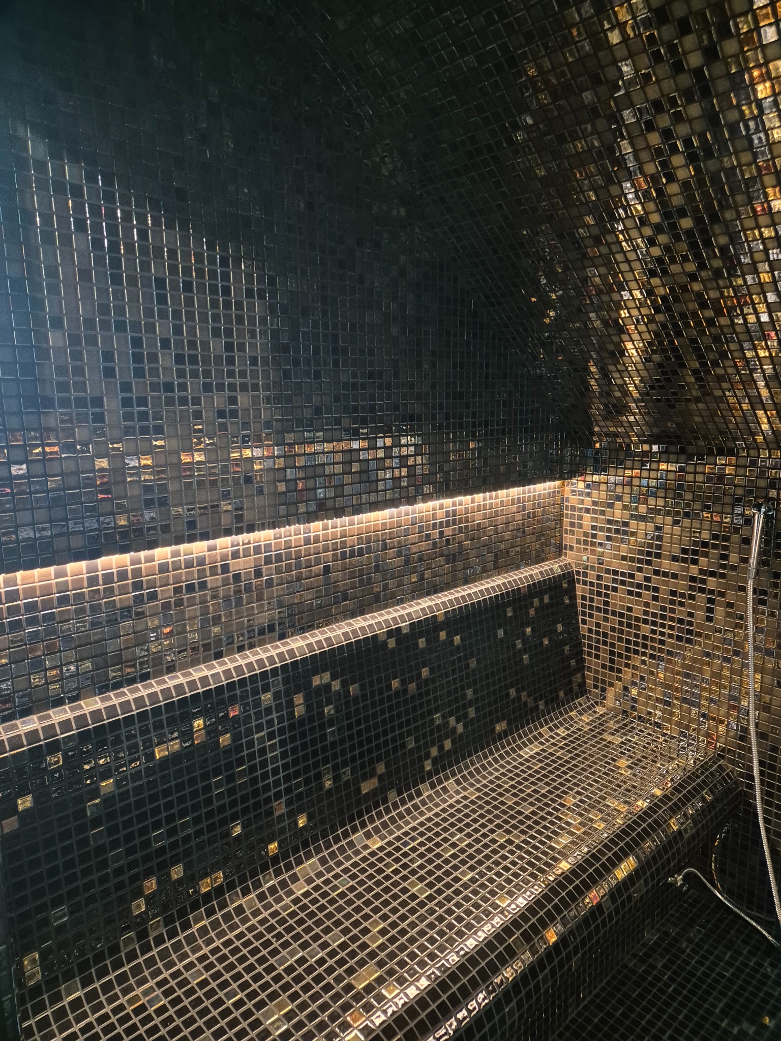 Wnętrze sauny wyłożone czarną i złotą mozaiką z podświetlanymi półkami i prysznicem.