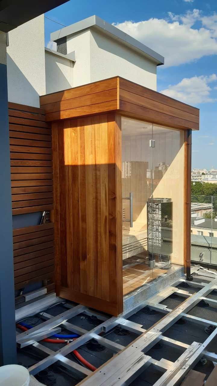 Drewniana sauna z przeszklonymi drzwiami na tarasie w trakcie montażu, widoczne elementy konstrukcyjne podłoża i instalacje.