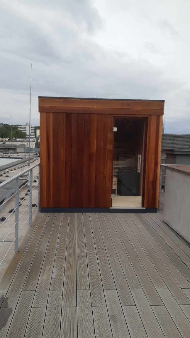 Sauna zewnętrzna z drewna egzotycznego, z przeszklonymi drzwiami, postawiona na tarasie drewnianym na dachu budynku w Czersku, widok z boku.