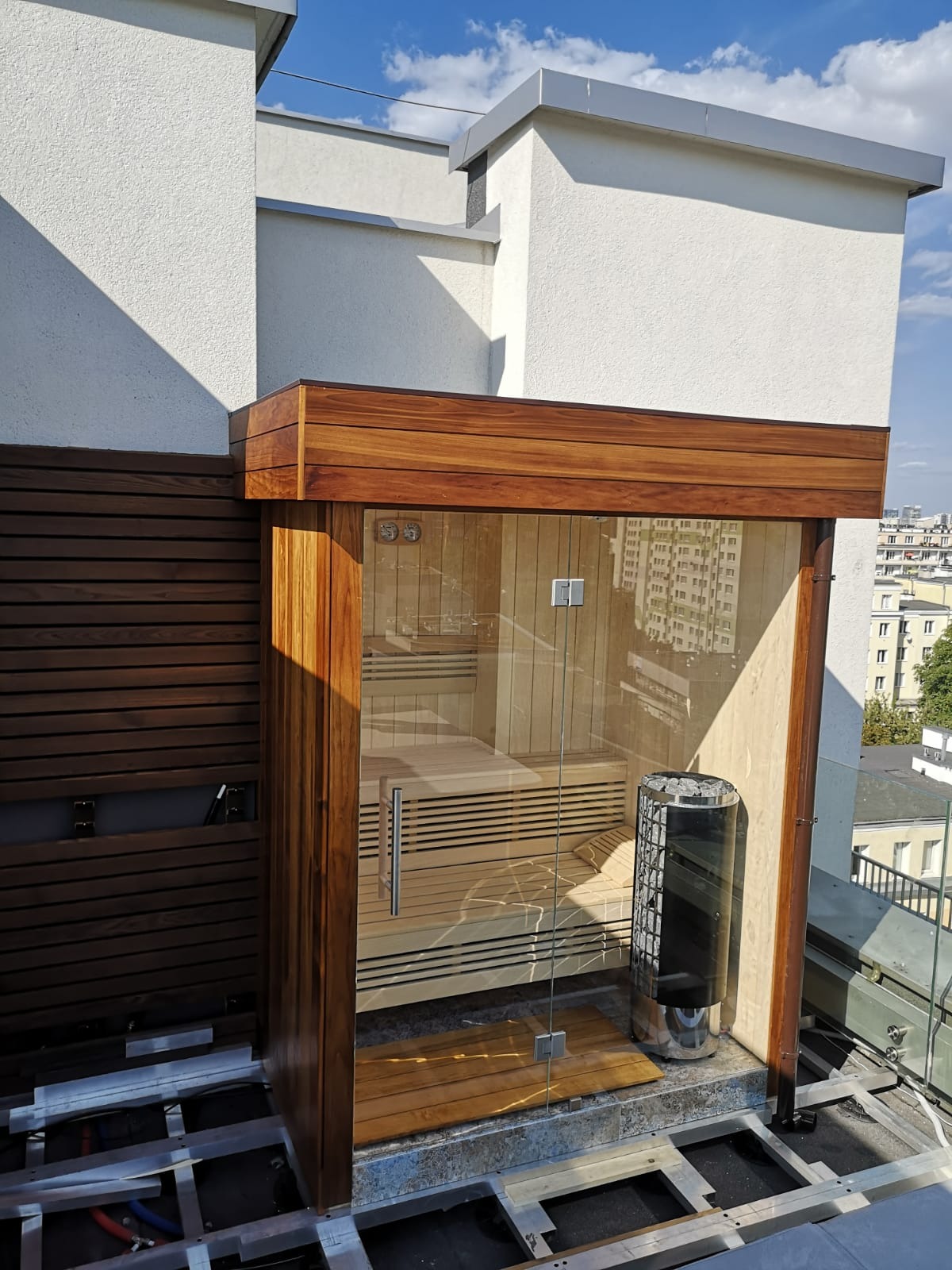 Zewnętrzna sauna na dachu budynku, z drewnianym wykończeniem i przeszklonymi drzwiami, widoczny piec i drewniane ławki wewnątrz, w tle panorama miasta.
