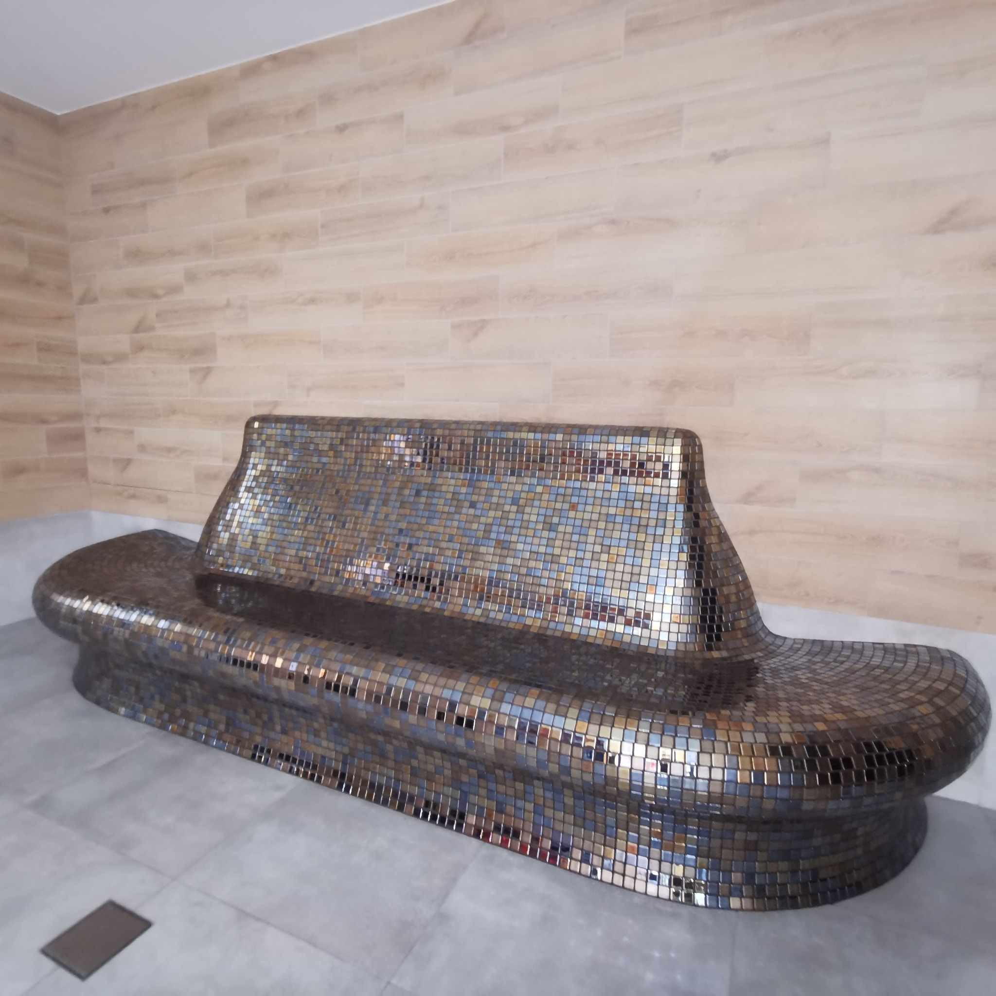 Mozaikowa ławka z oparciem w strefie relaksu, wykończona drobnymi, połyskującymi płytkami w odcieniach brązu i szarości, na tle ściany z płytek imitujących drewno.