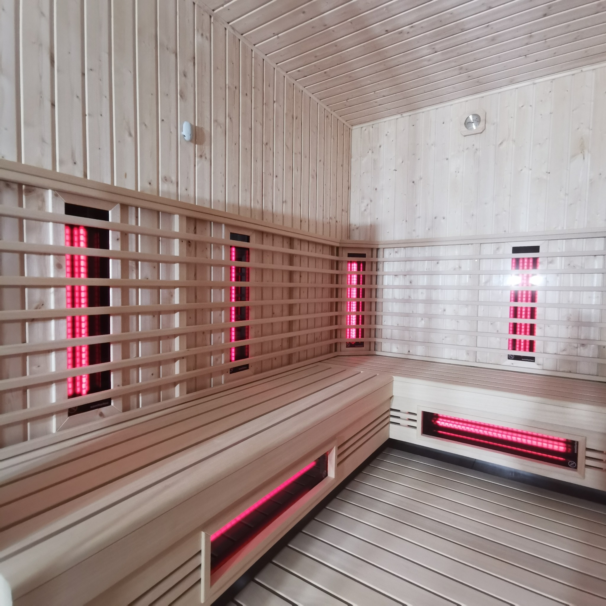 Wnętrze sauny infrared z jasnego drewna, z ławkami i panelami grzewczymi emitującymi czerwone światło, oświetlenie sufitowe.