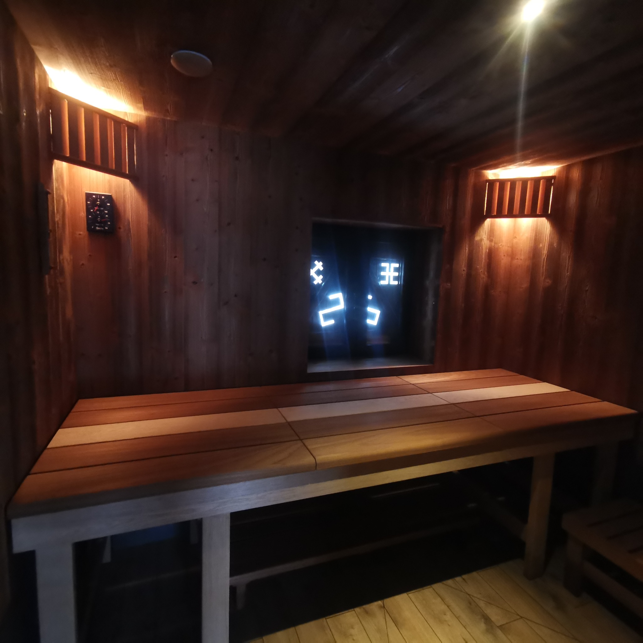 Wnętrze sauny z drewnianymi panelami, ławami z jasnego drewna i wyświetlaczem temperatury w tle.