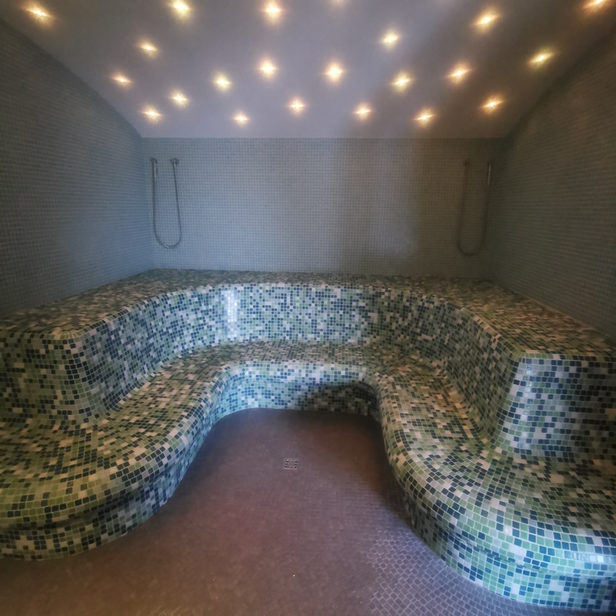 Wnętrze łaźni parowej z mozaikową ławą w odcieniach zieleni i błękitu, oświetlone punktowymi lampami sufitowymi, z odpływem podłogowym i prysznicami na ścianach.