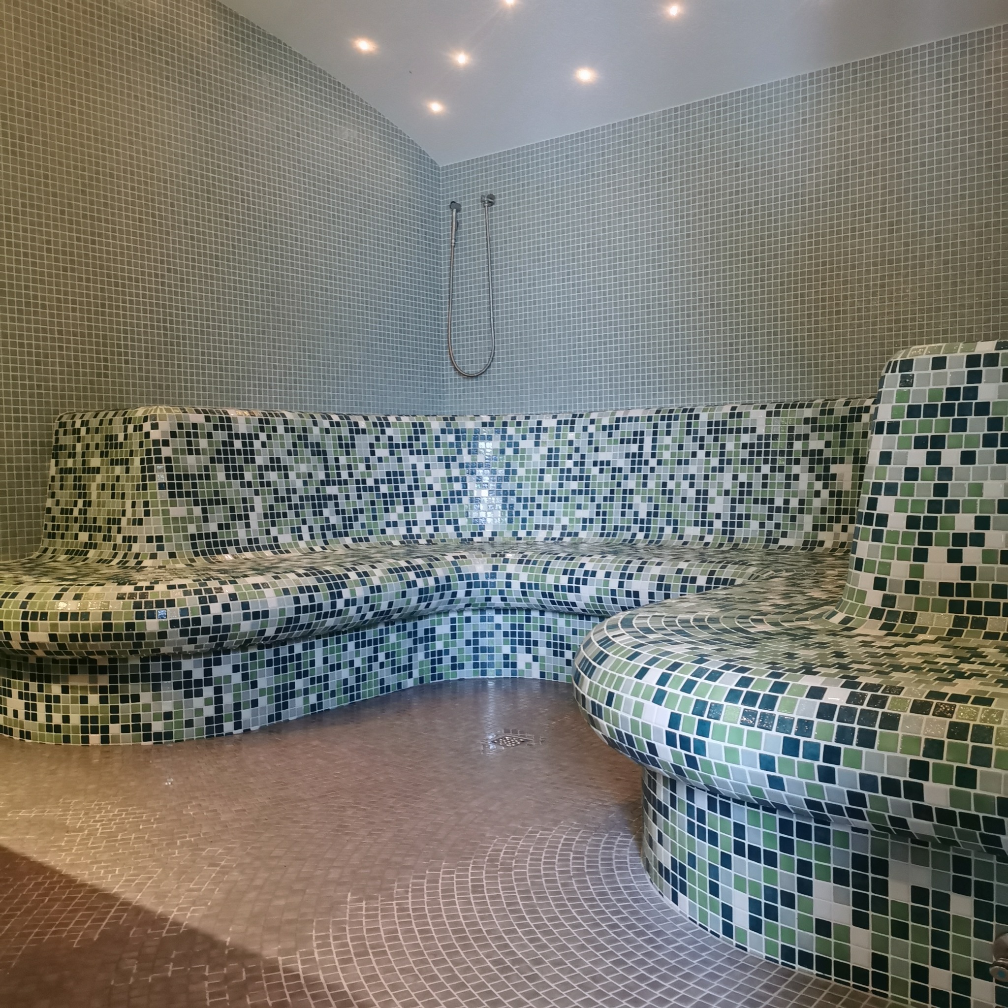 Wnętrze sauny wyłożone mozaiką w odcieniach zieleni i błękitu, z ławkami o zaokrąglonych kształtach i prysznicem w rogu.